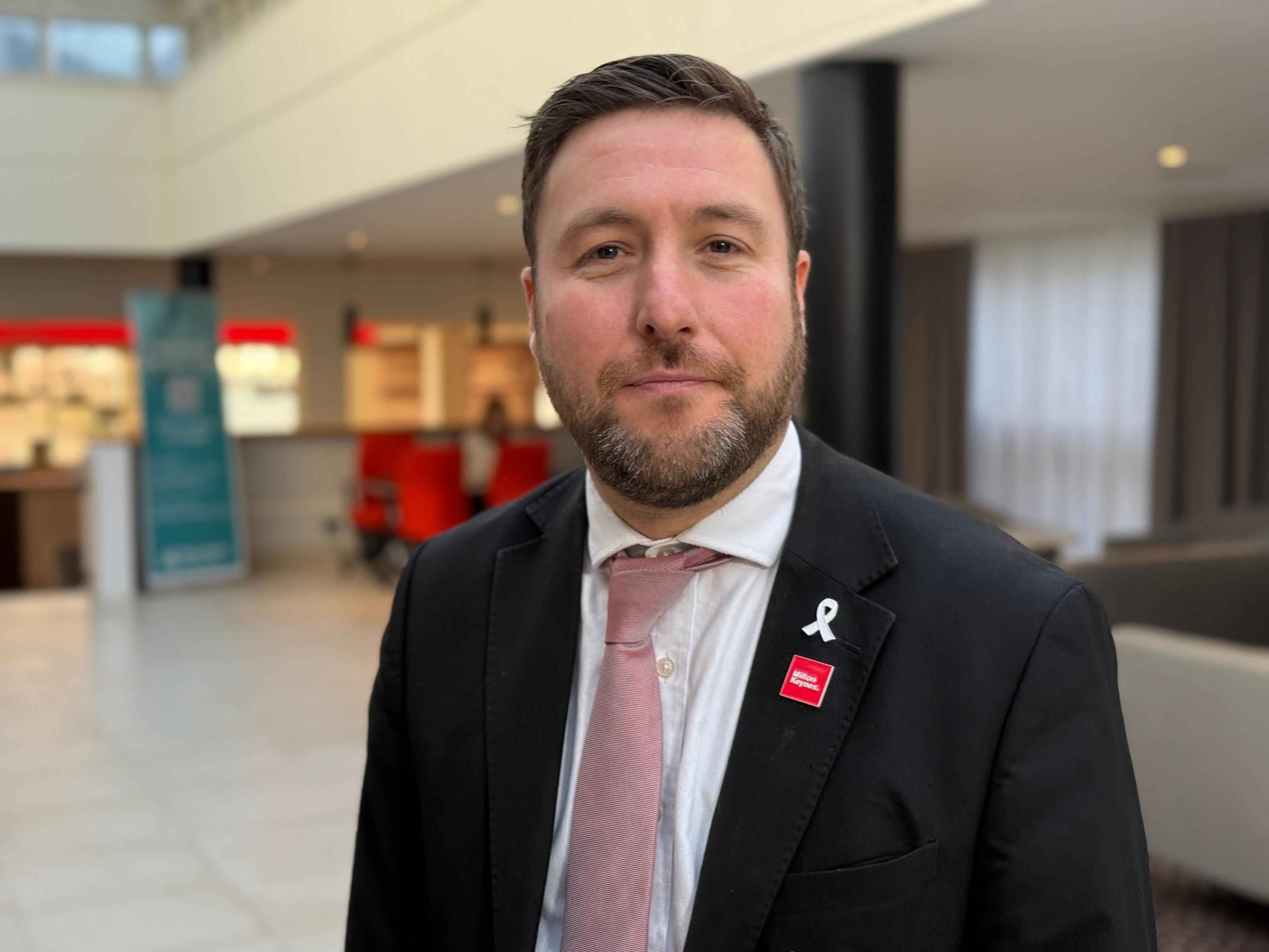 A picture of a man who has short dark brown hair and a dark brown beard with white bits. He is wearing a black suit jacket with a dark pink tie.