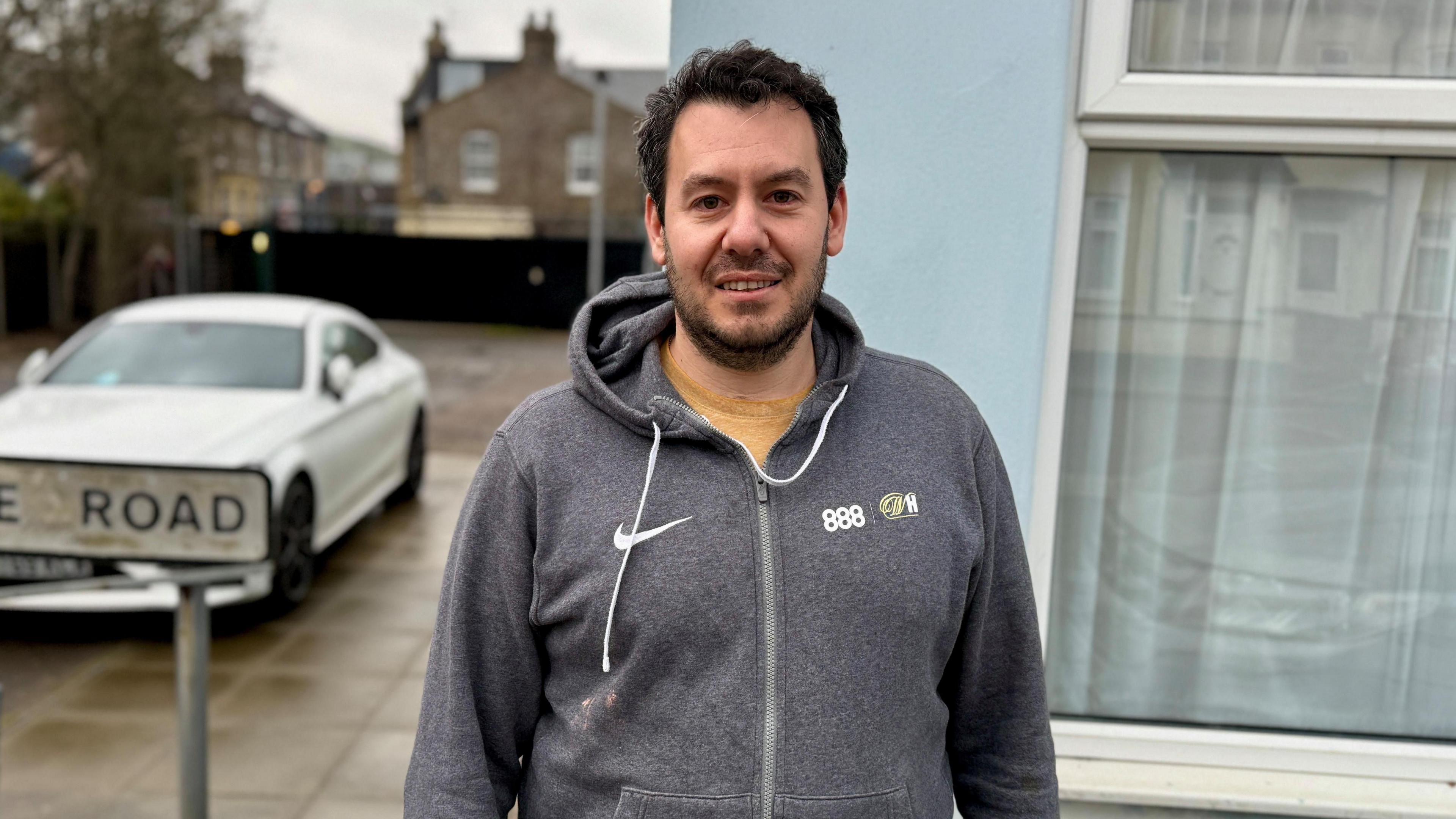 Darren Bloch is wearing a grey hoody and has short brown hair with a brown beard.  His is stood outside his house which is blue, you can see the road that runs alongside it towards the EastEnders set