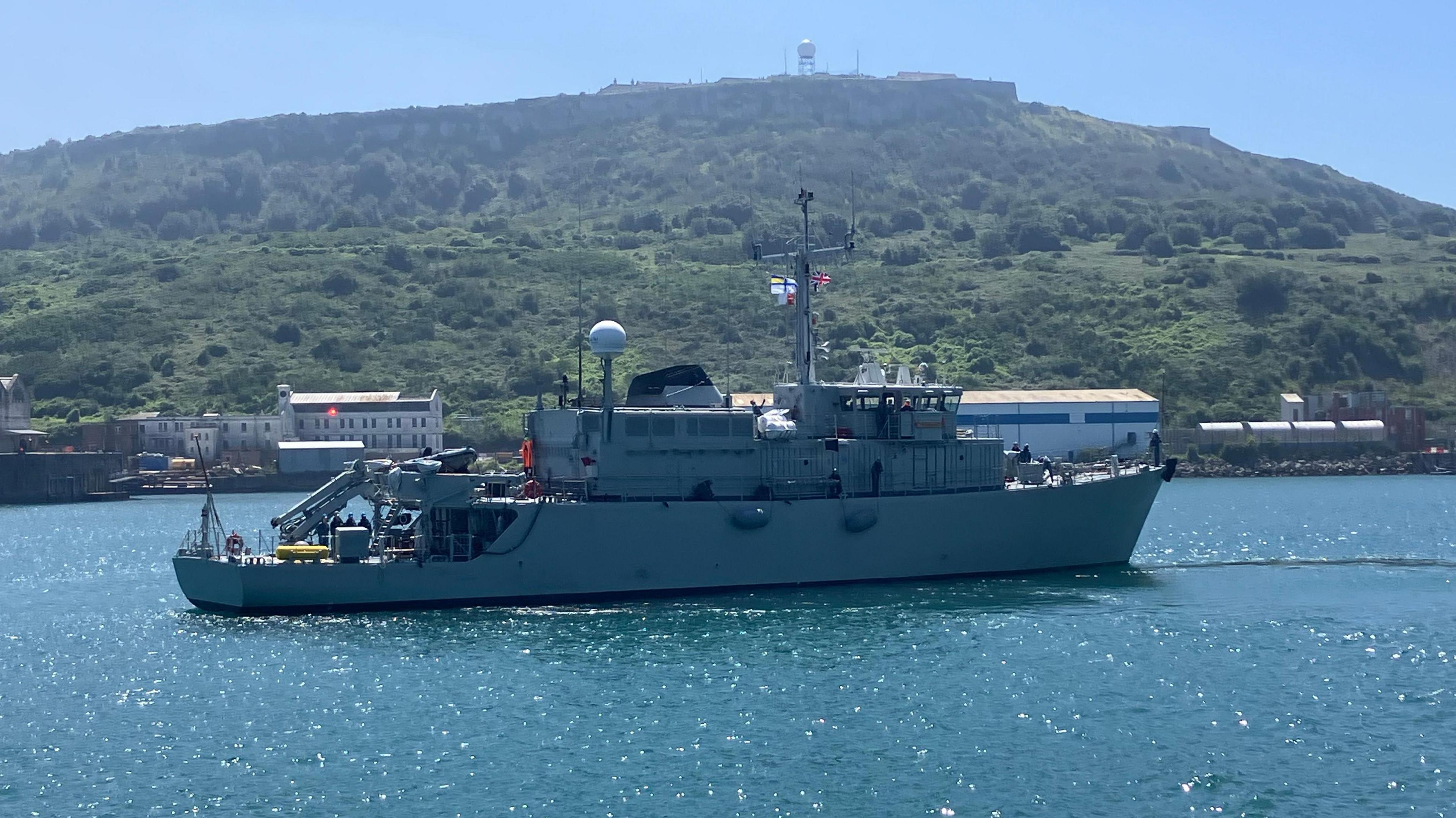 A warship on the water sailing to the right with a union jack visible and a Ukraine Navy flag also visible with some green hills in the background