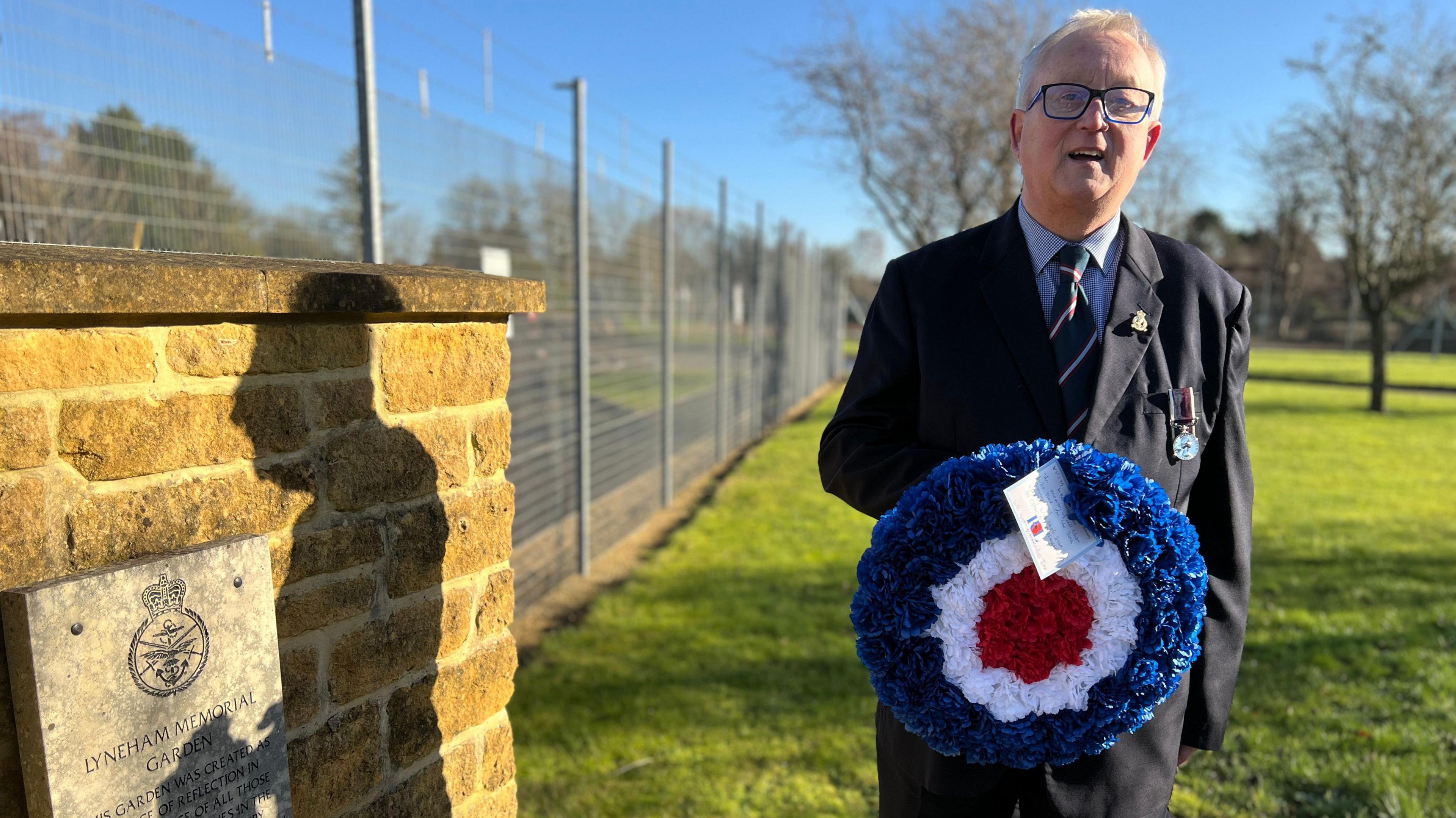 Geoff Bridgman at the REME remembrance garden in Lyneham. He is standing next to a brick wall with a plaque on it. Geoff has short white hair, glasses and is wearing a black suit and striped tie. He is holding a red white and blue circle wreath. He is also looking at the camera and smiling 