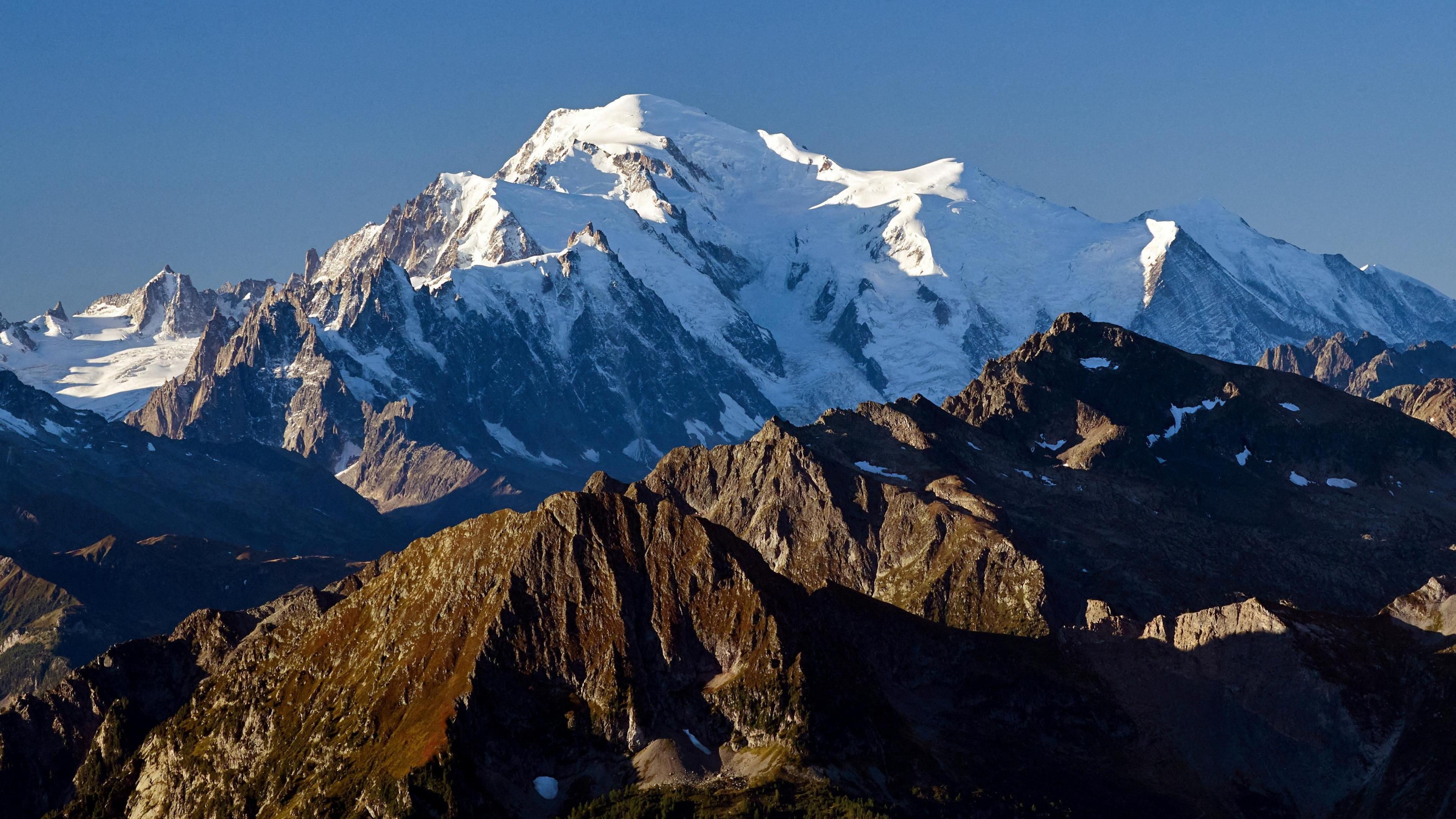 A file photo of Mont Blanc. A brown mountain stands in front of a snow-covered mountain under a blue sky.