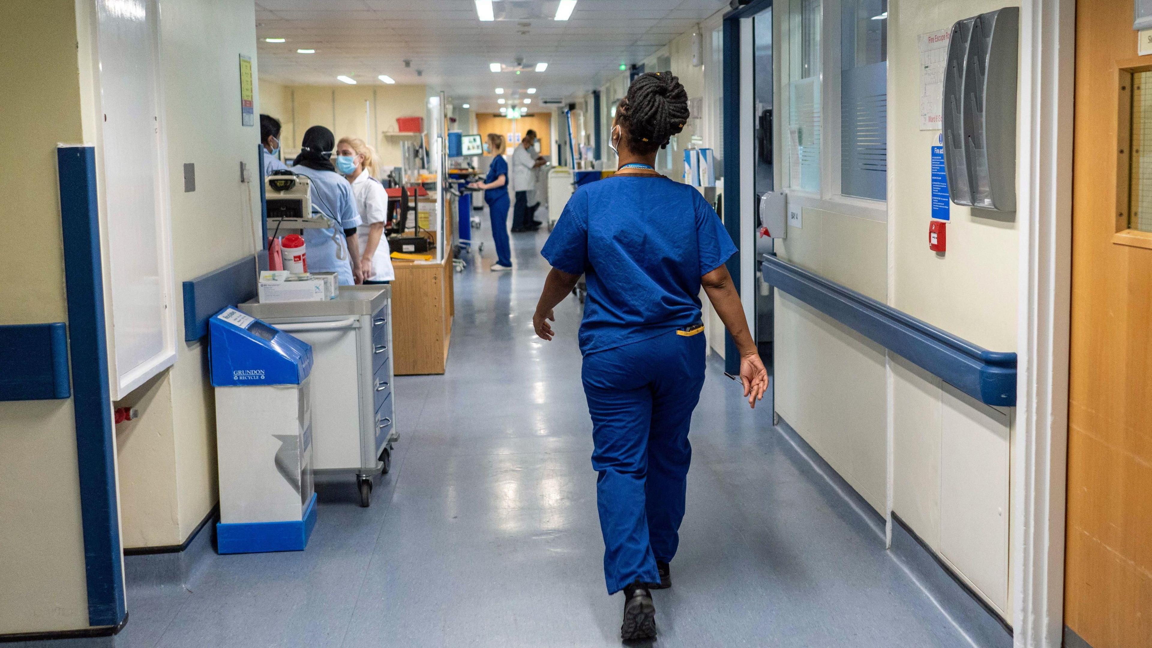 General view of a hospital ward - a medical professional walks down the middle- with other staff in masks on the left 