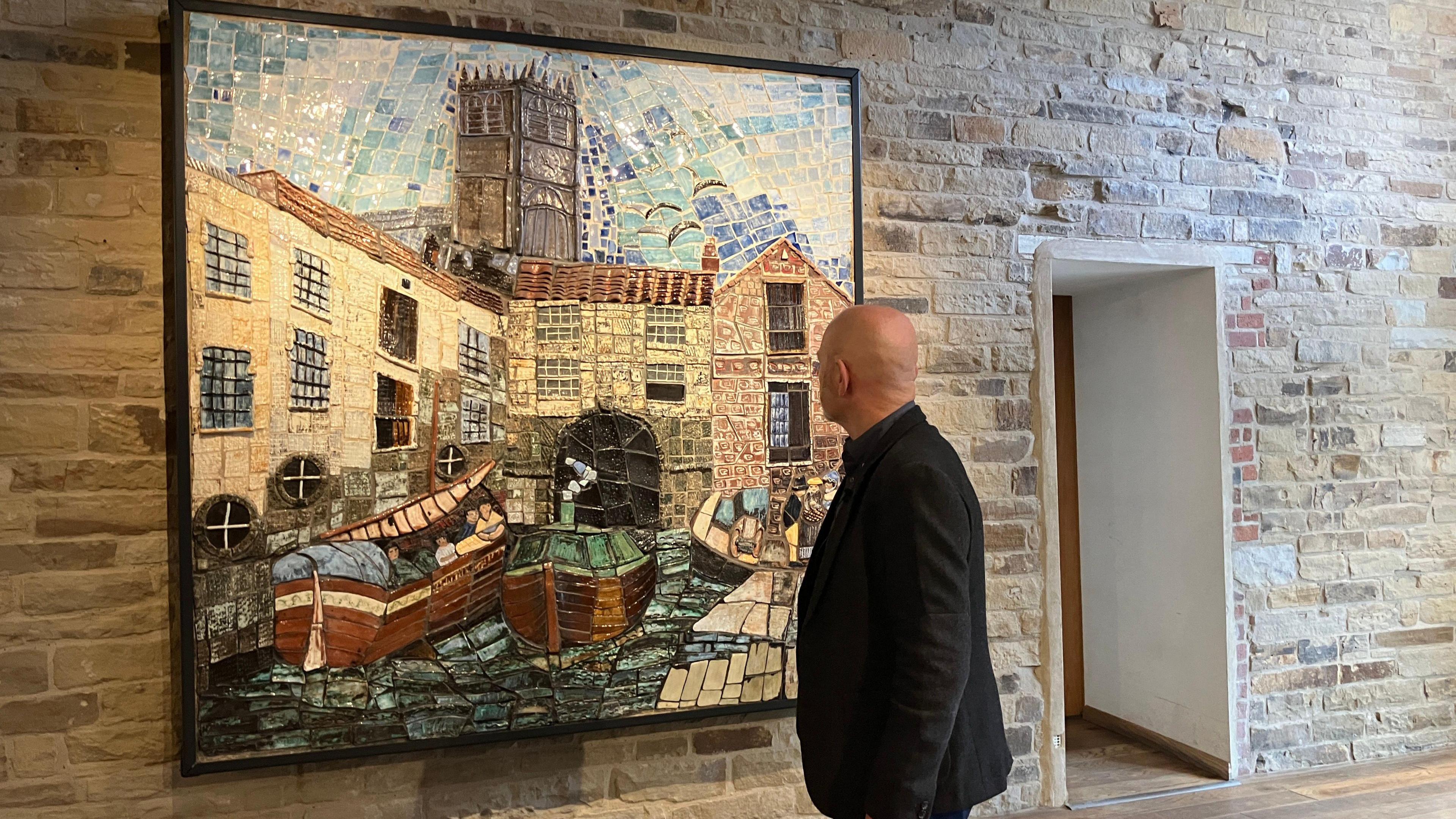 A man, wearing a black blazer, looks at a multi-coloured mural hanging on a stone wall