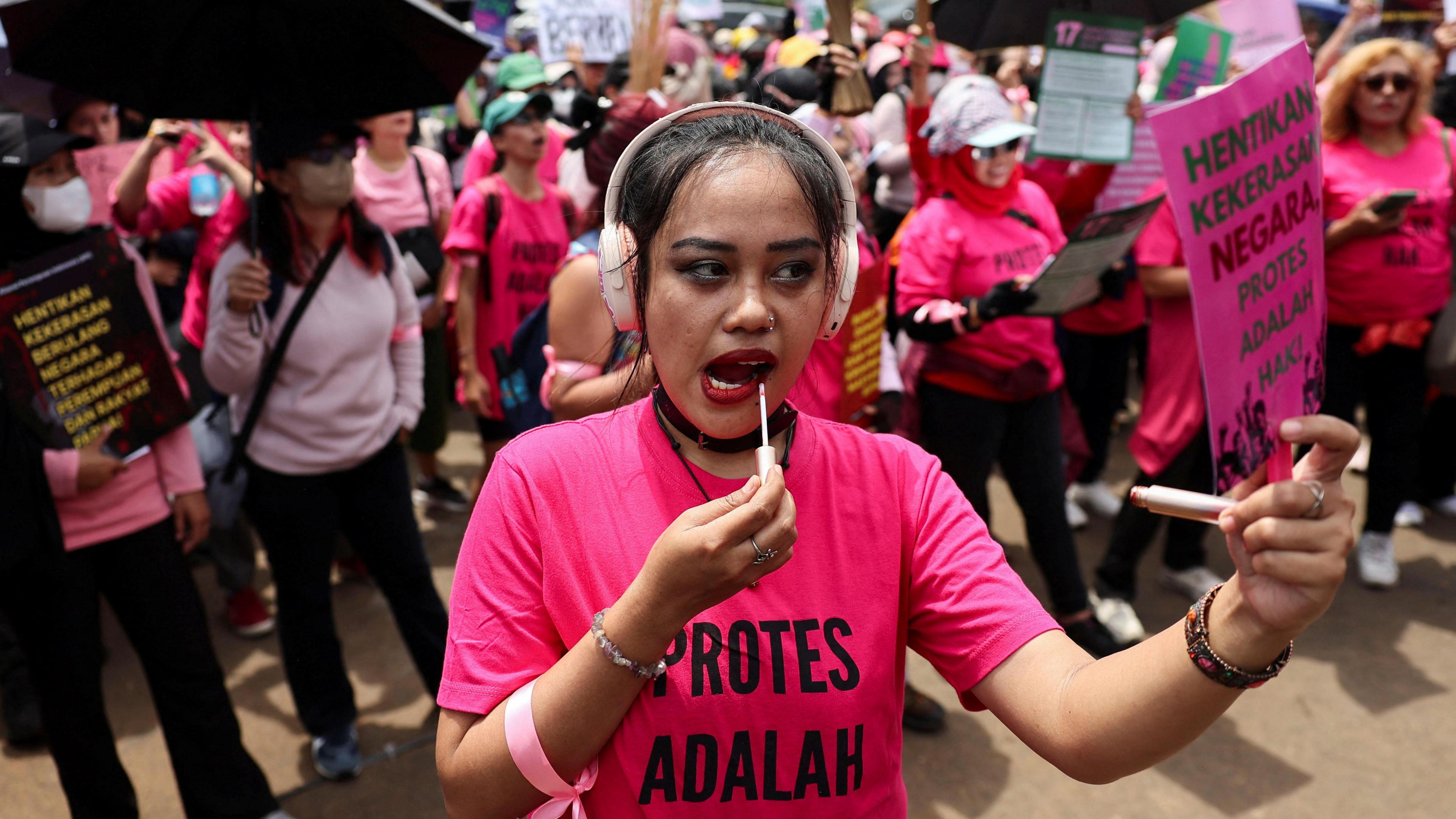 A woman wearing a pink shirt and pink headphones puts on lipstick while holding a pink sign during a protest in Indonesia