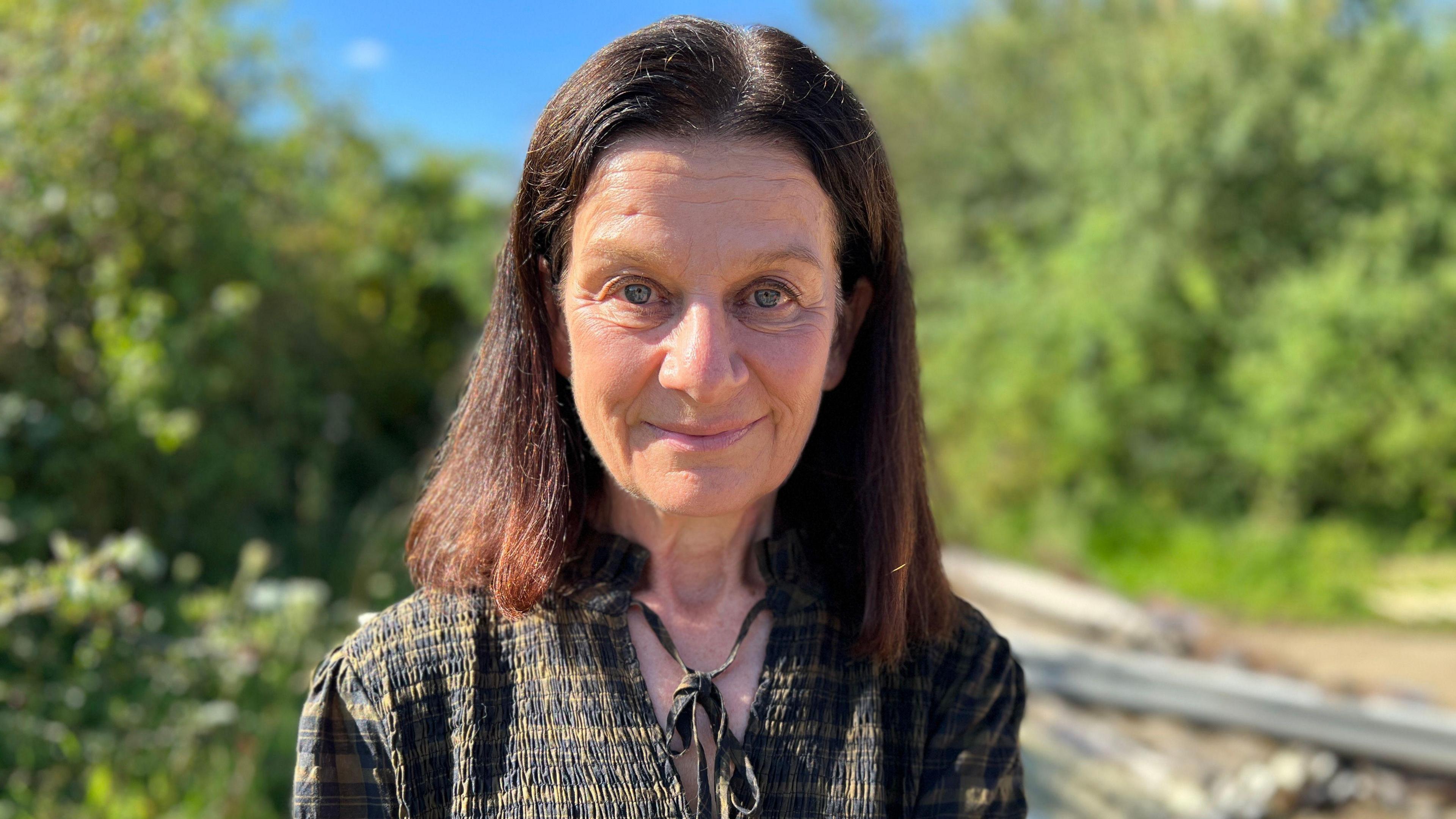 Bridget Smith is looking at the camera. She has long brown hair and she is wearing a red-and-brown open-necked top. In the background there are fields and hedges. It is a sunny day.