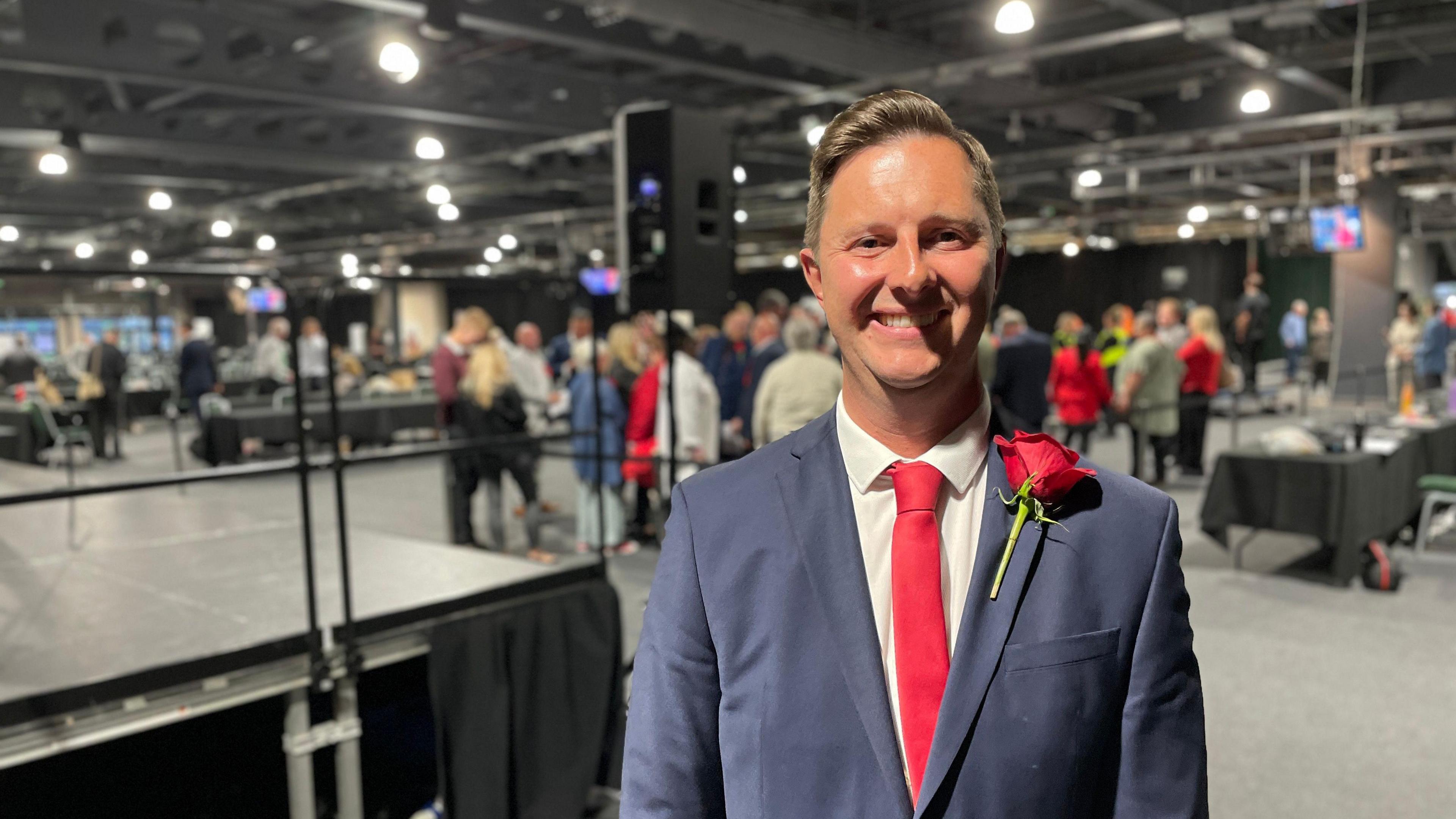 A smiling man wearing a suit and tie looks at the camera. He is in a hall with people and display tables behind him.