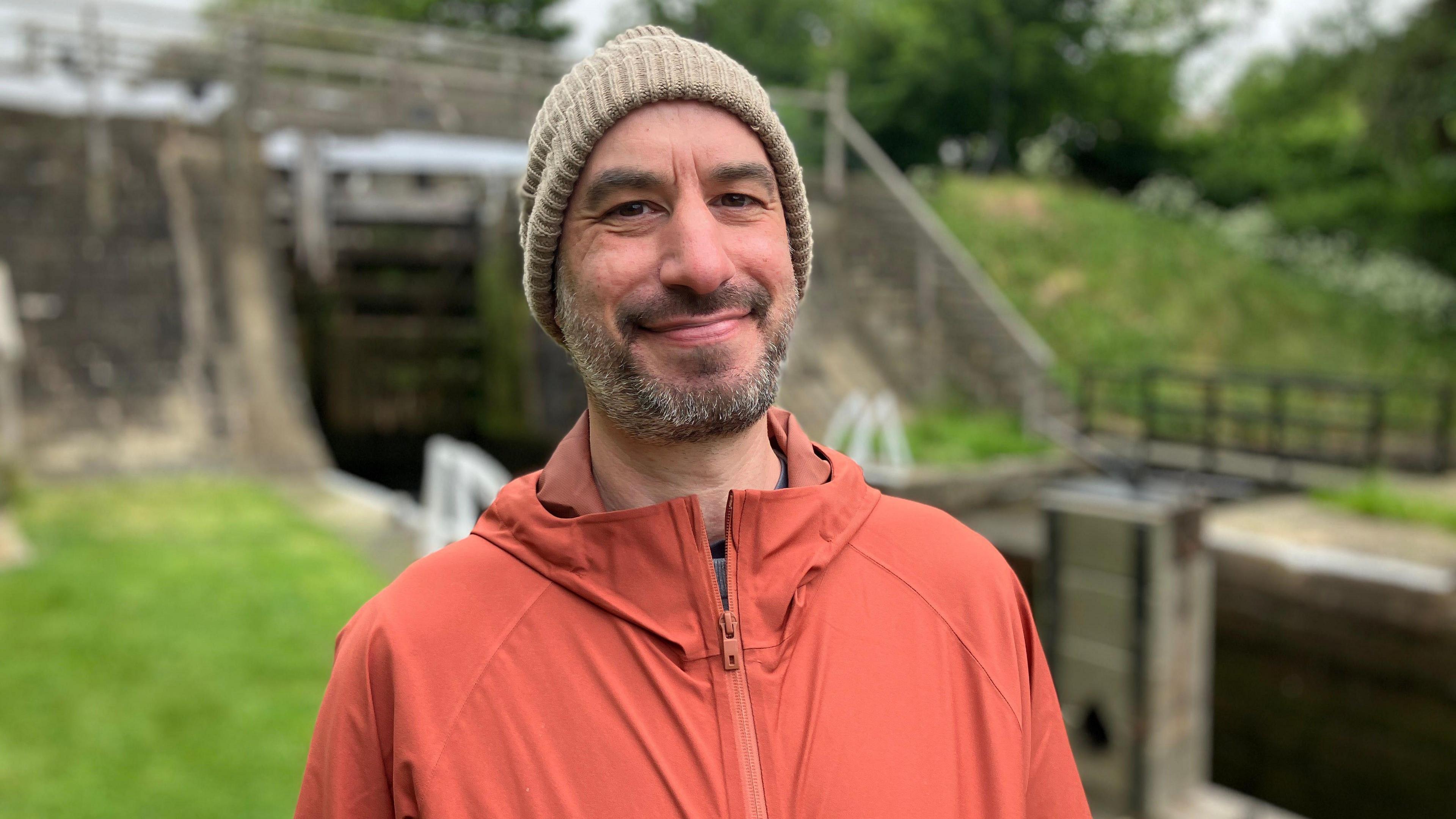 A man stands in front of a canal lock. He is wearing an orange coat and hat as he smiles at the camera.