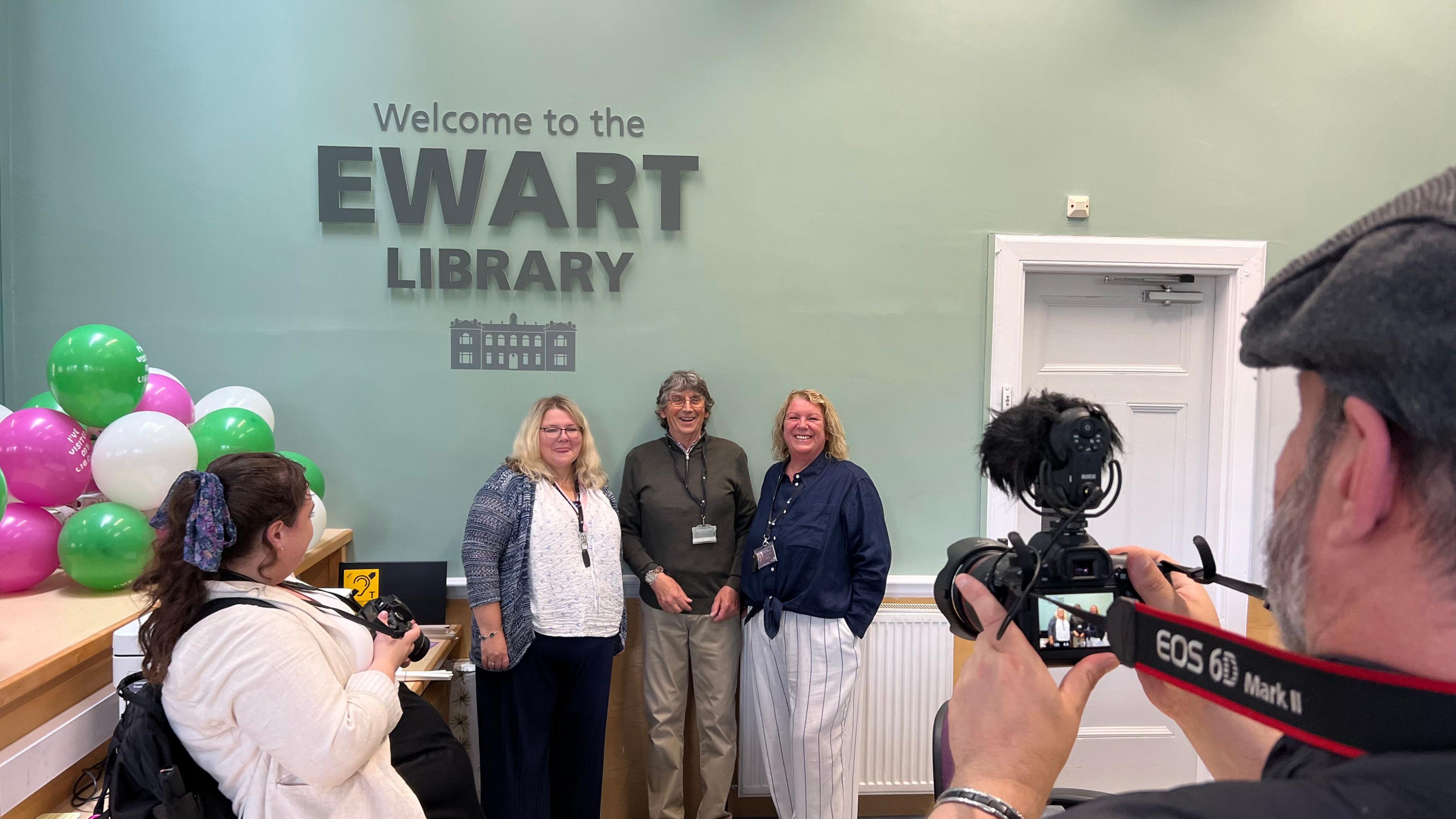 Over the shoulder picture of a grey-ing bearded photographer taking a picture of three staff members with landyards underneath the welcome to the ewart library sign.