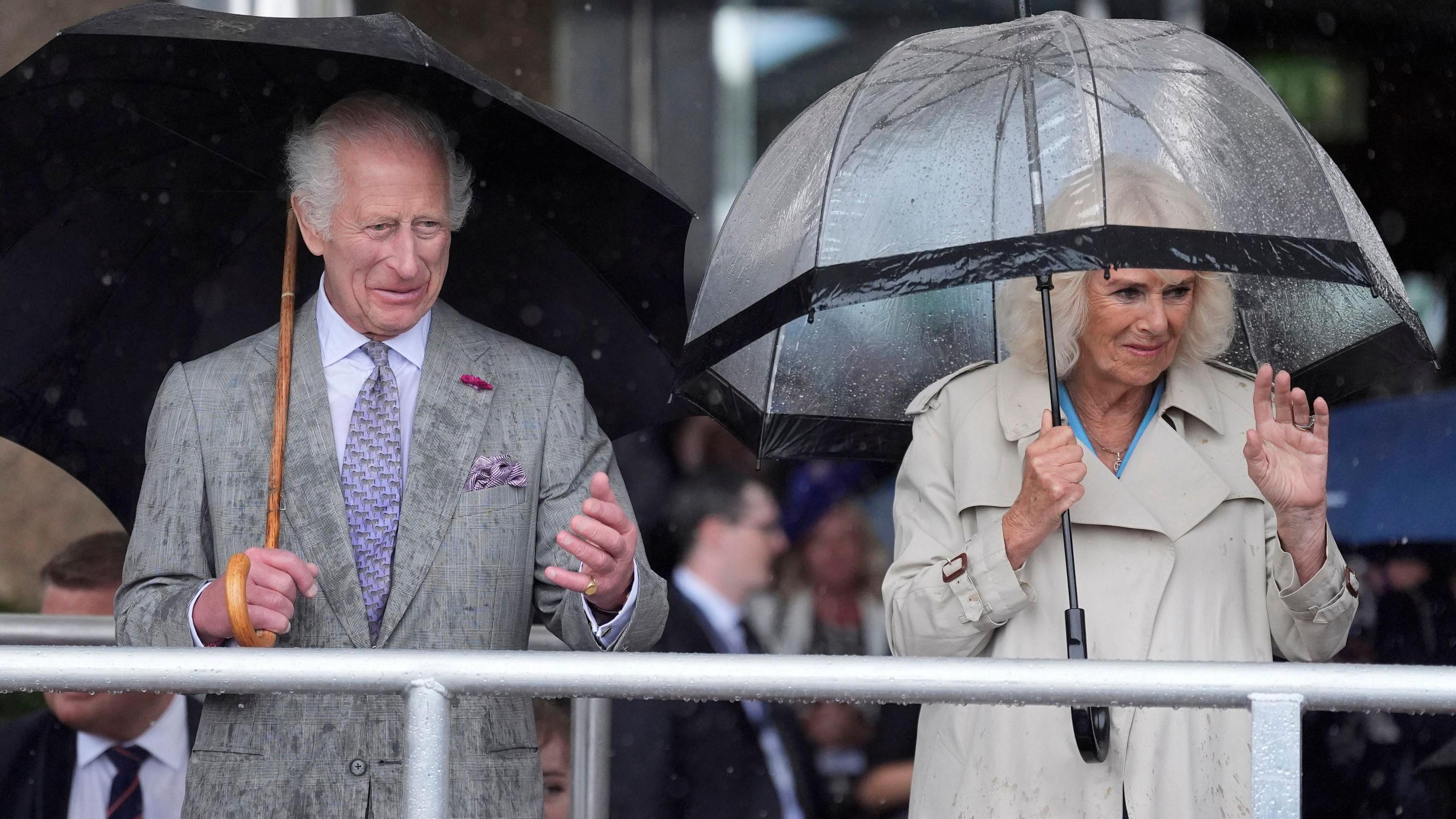 King Charles III (left) and Queen Camilla during the King's Parade at Liberation Square in St Helier, Jersey, during their two-day visit to the Channel Islands. The King is wearing a grey suit and the Queen is wearing a beige overcoat. Both are holding umbrellas above their heads. 