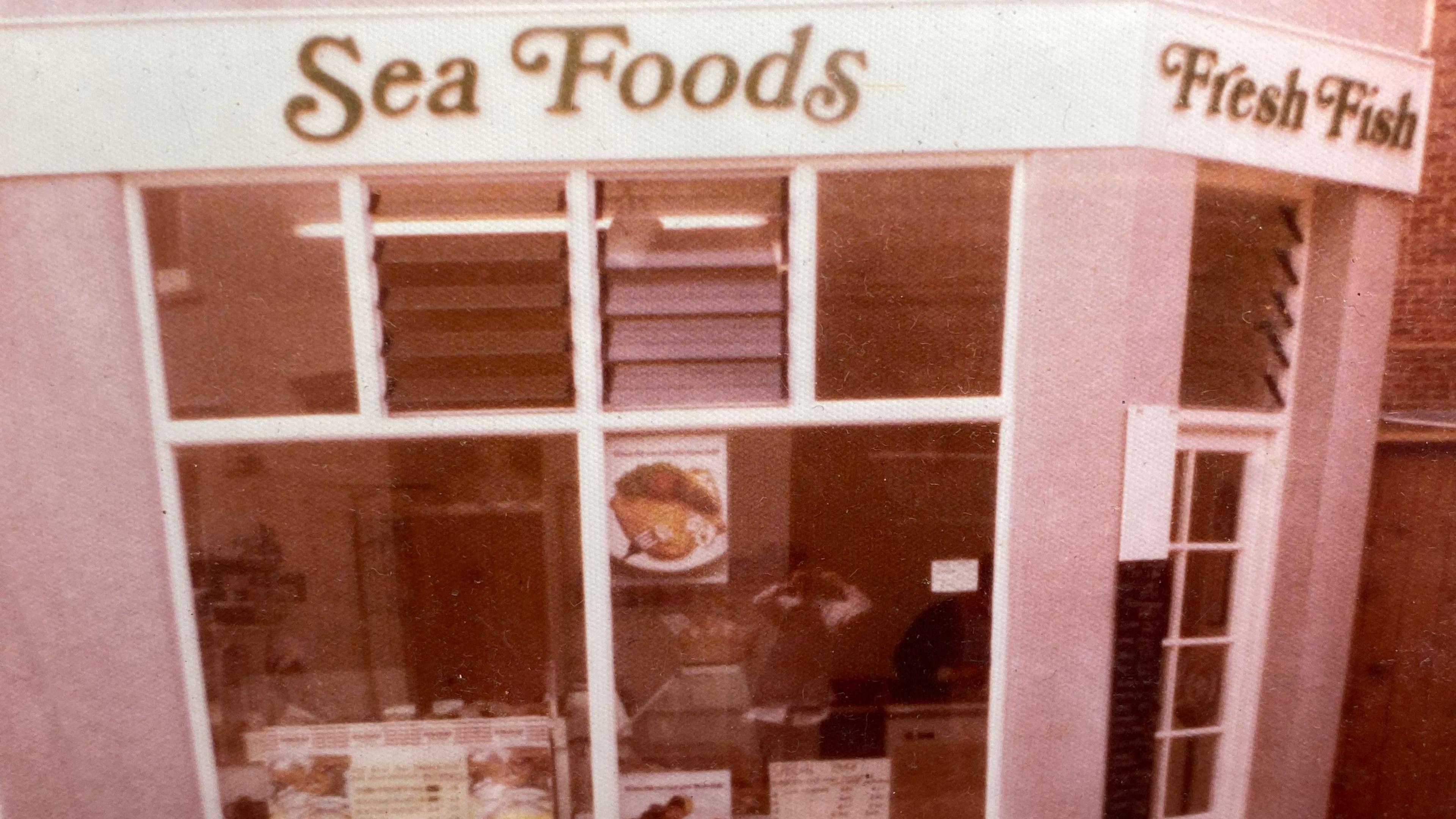 The front of the Sea Foods shop in the 1970s. It is a faded and reduced colour image with the reflection of the photographer on the window.
You can see a picture of fish in the window and a faded view of the counter inside.
Over the door to the right of the picture, fresh fish is written in italic.