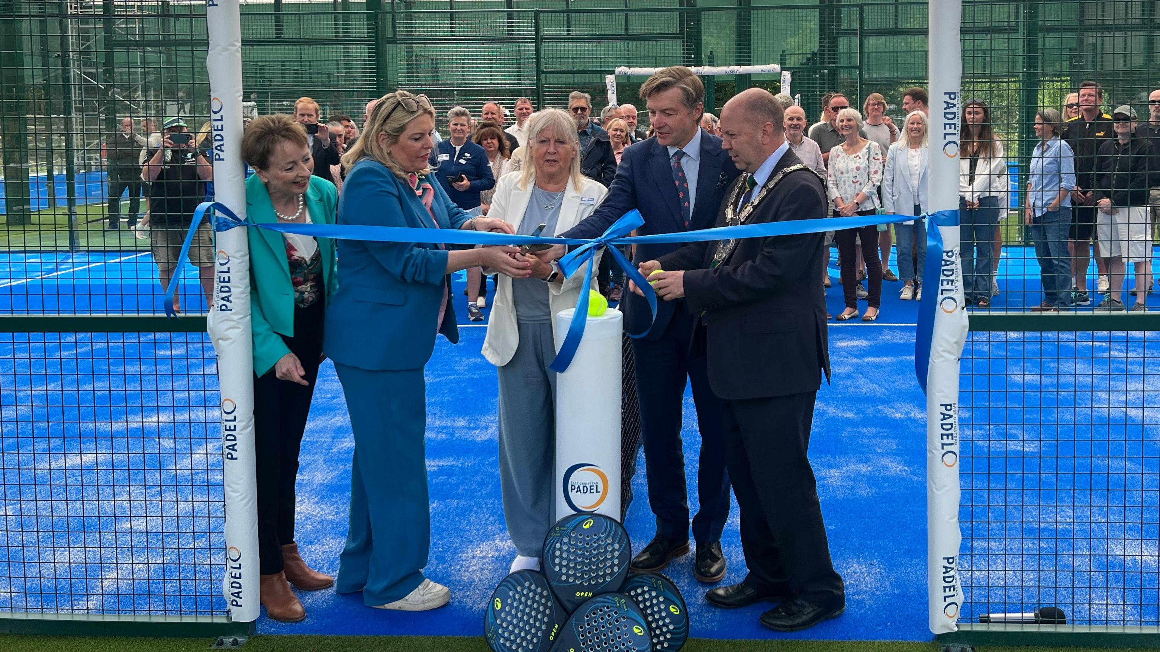 Five people pictured at the opening of the new padel facility in East Grinstead, West Sussex. A lady in the middle wearing a white jacket can be seen cutting a blue ribbon.