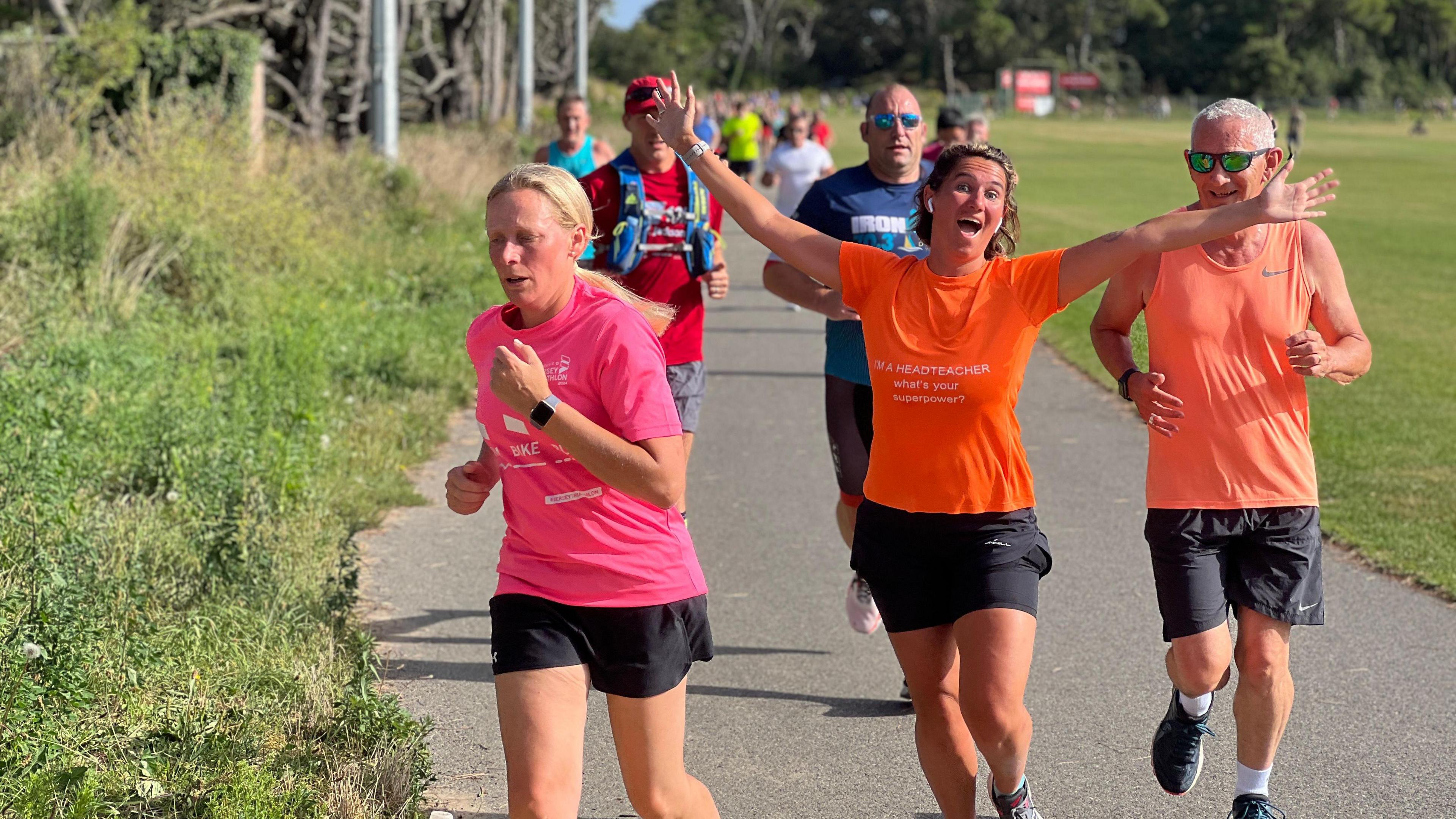 A runner smiles at the camera with her arms spread wide as she runs with a group of people