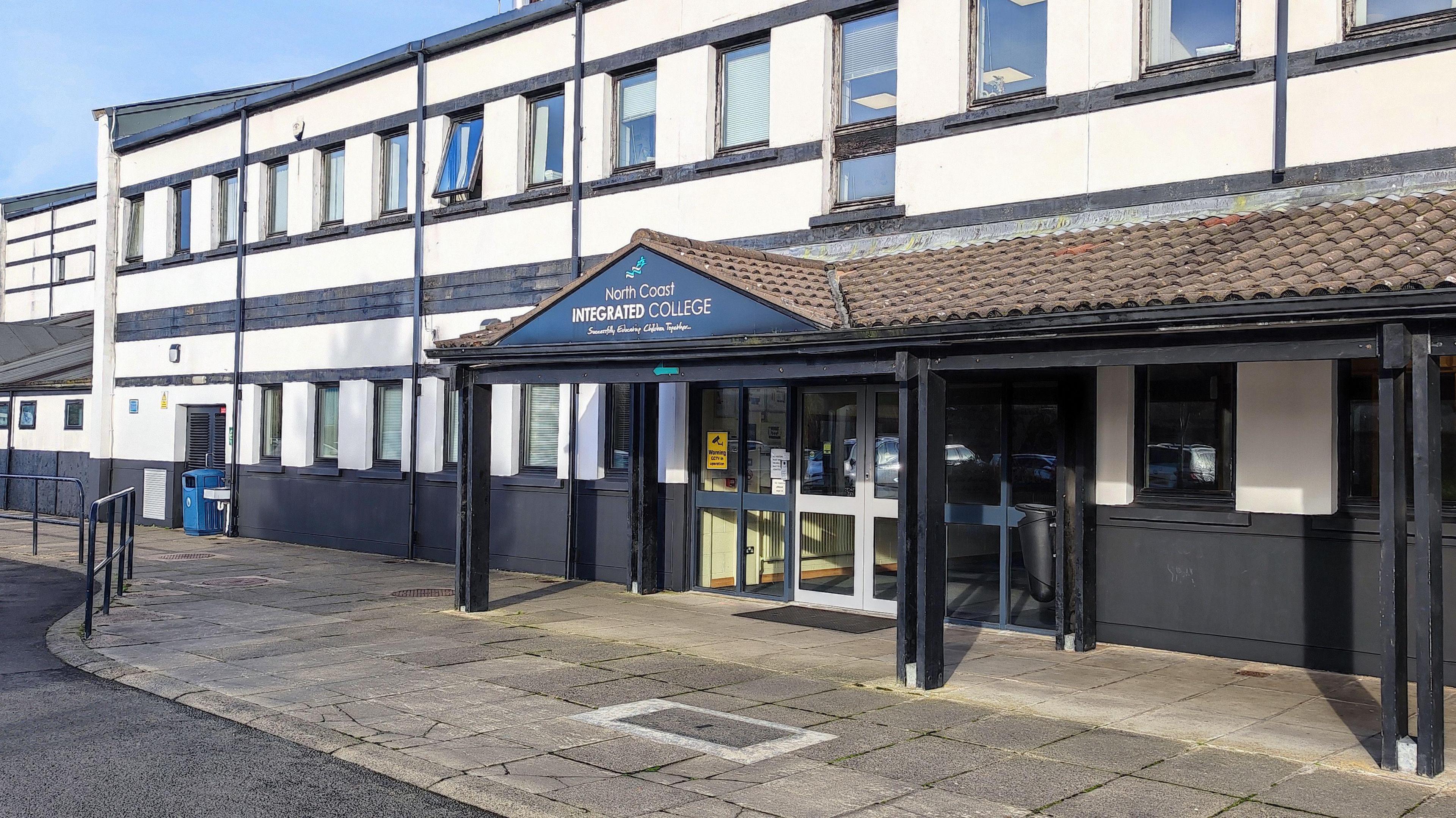An exterior photo of a white building with black trim. There is a sheltered area above the door, the roof is brown and on a black sign it says, North Coast Integrated College.