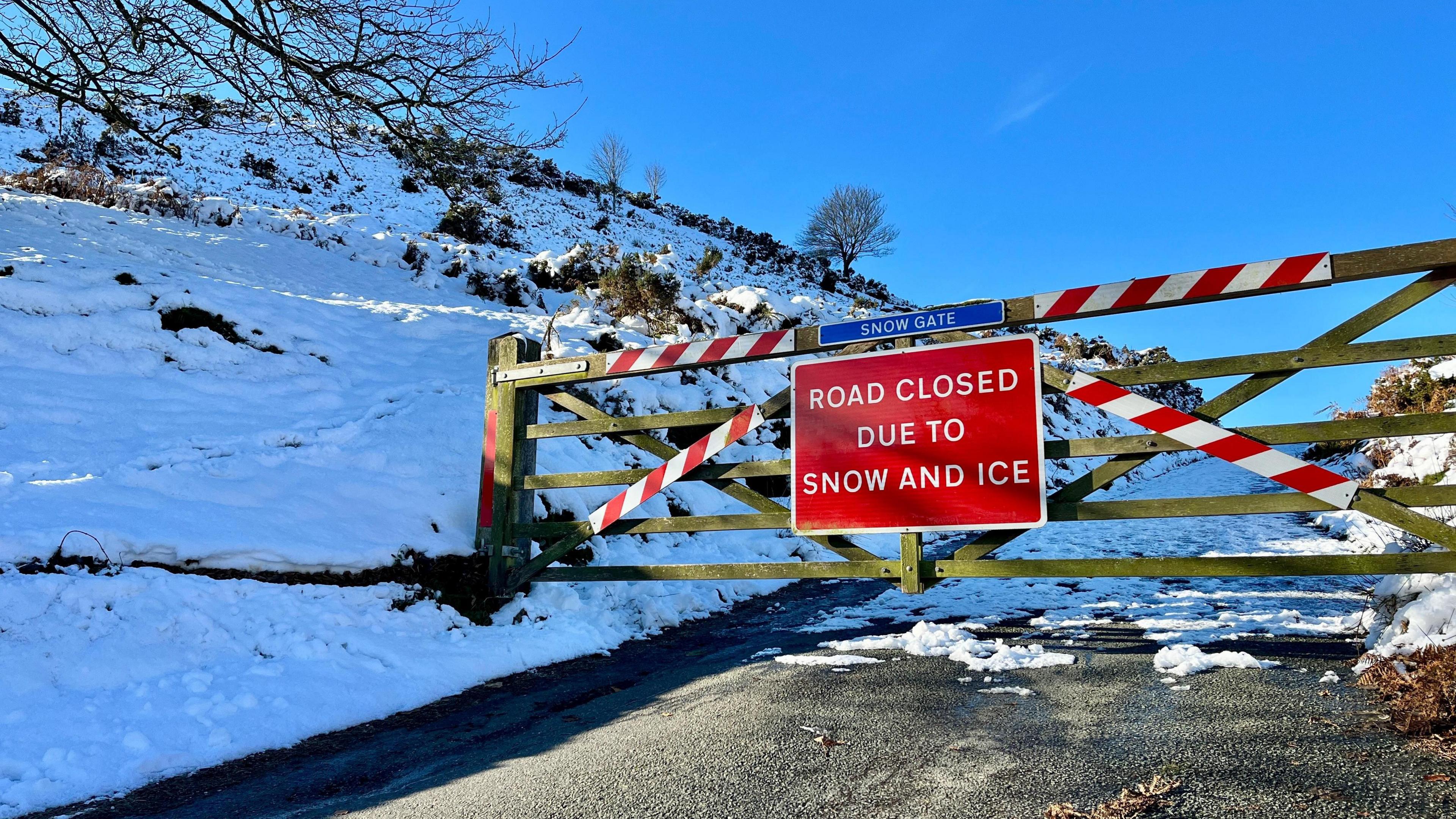 A five-bar wooden gate across a road with a road sign "Road closed due to snow and ice". A snowy landscape lies behind, with compressed snow on the carriageway. 