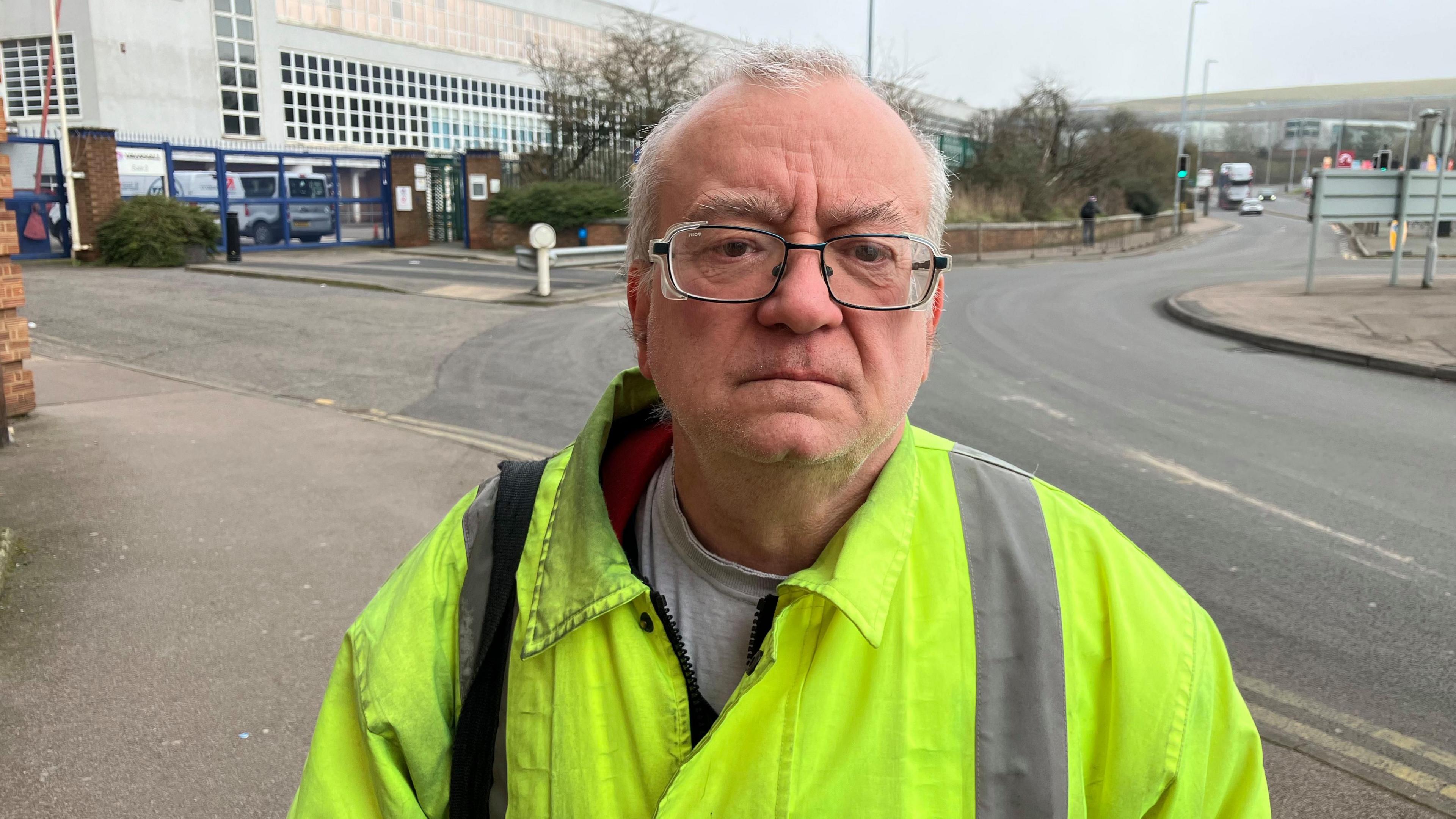 Adrian is wearing a high vis jacket with a grey t-shirt underneath. He has black rimmed glasses and grey receding hair. He stands outside the Vauxhall factory next to the roundabout on Kimpton Road. His expression is serious with a slightly furrowed brow.