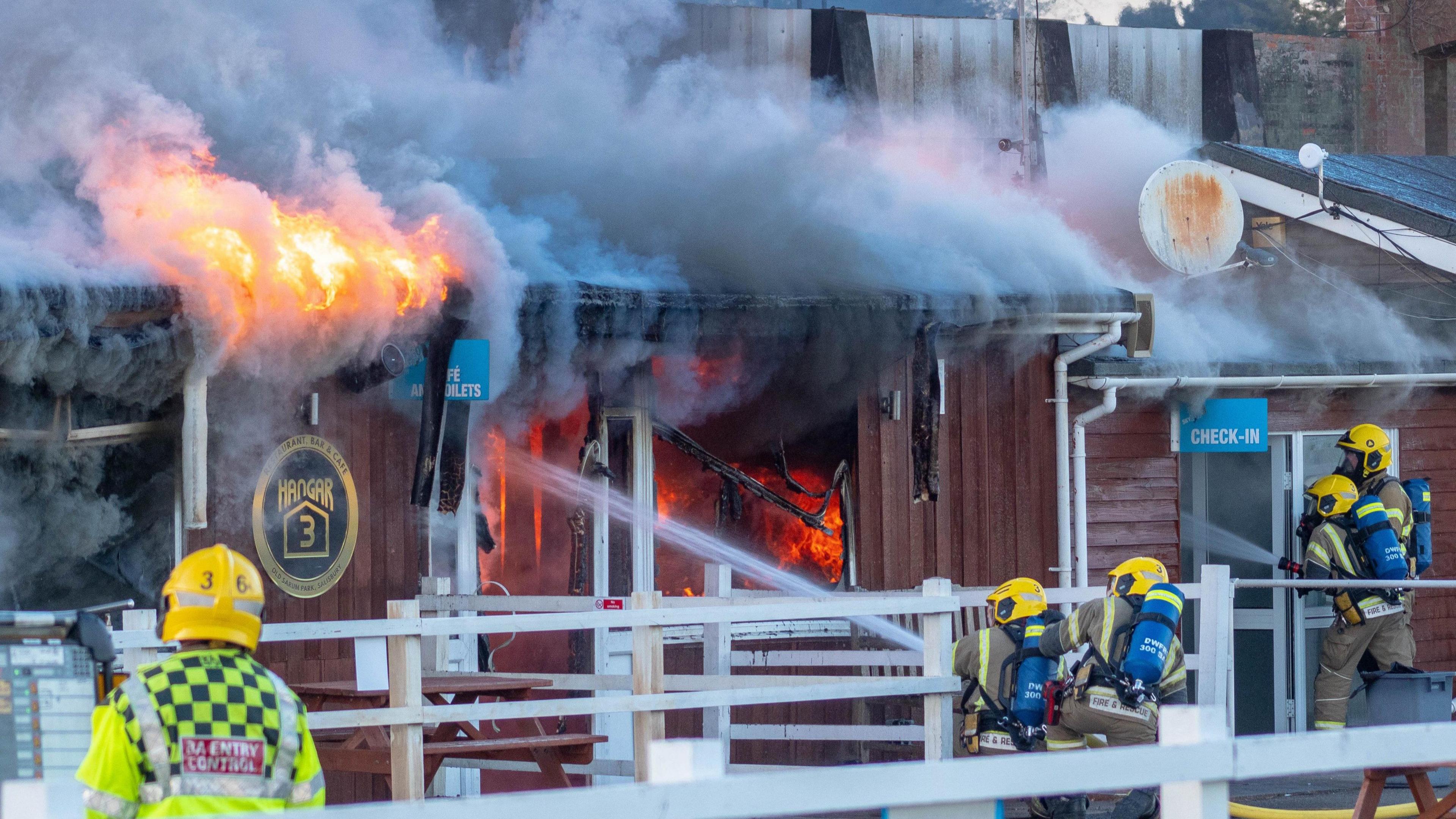 Firefighters are tackling a blaze at an airfield hangar. Firefighters wearing protective clothing are shooting water into an empty wooden hangar which is well alight. There is smoke coming off the roof.