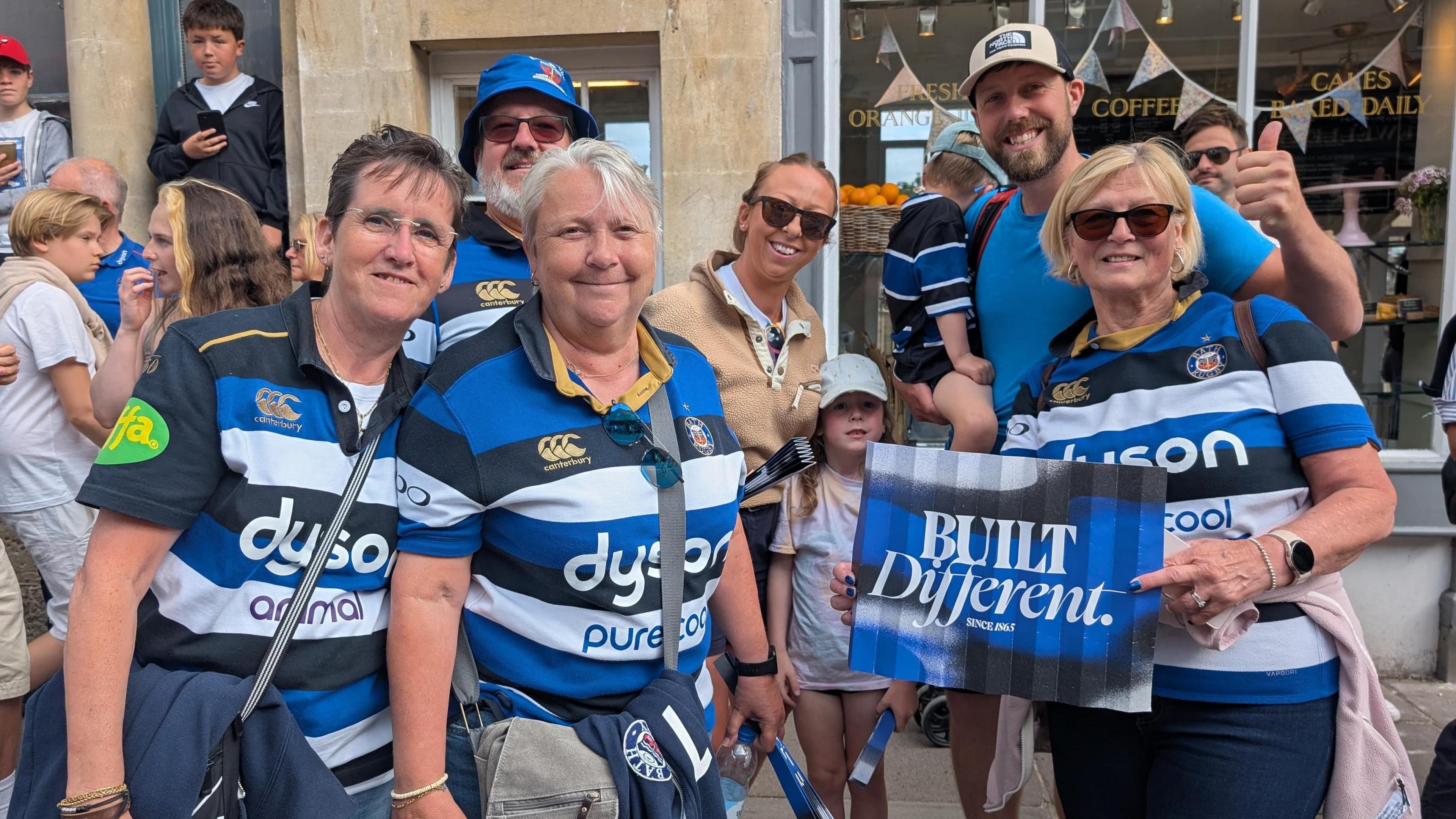 Seven people, five of whom are in Bath shirts for the parade. They are standing in front of a coffee shop with coloured bunting in the window