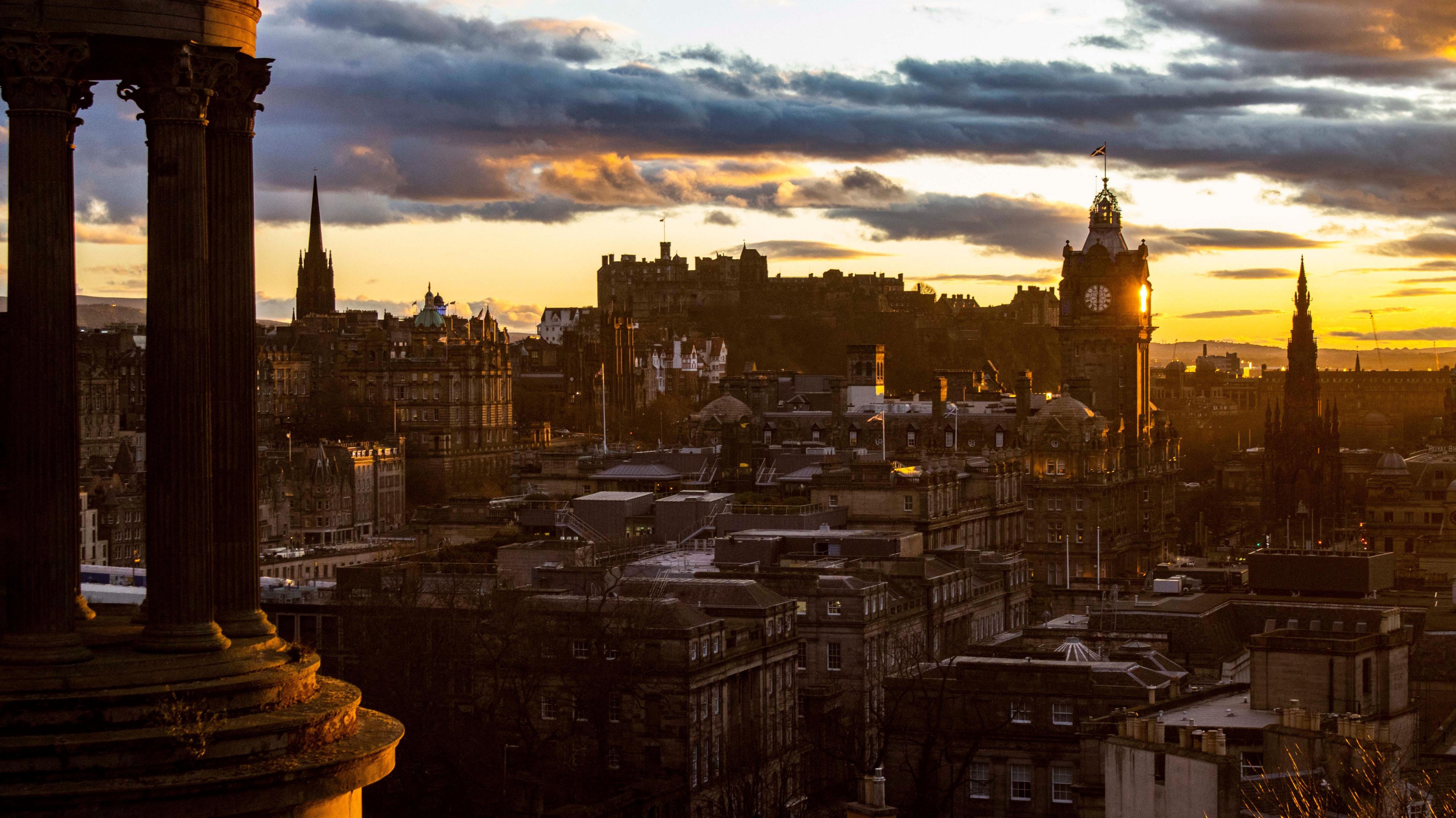 A general view of Edinburgh's New Town from Calton Hill. A monument is on the left of the picture. The new town can be seen below at sunset. The sky is orange with streaks of blue-grey cloud.