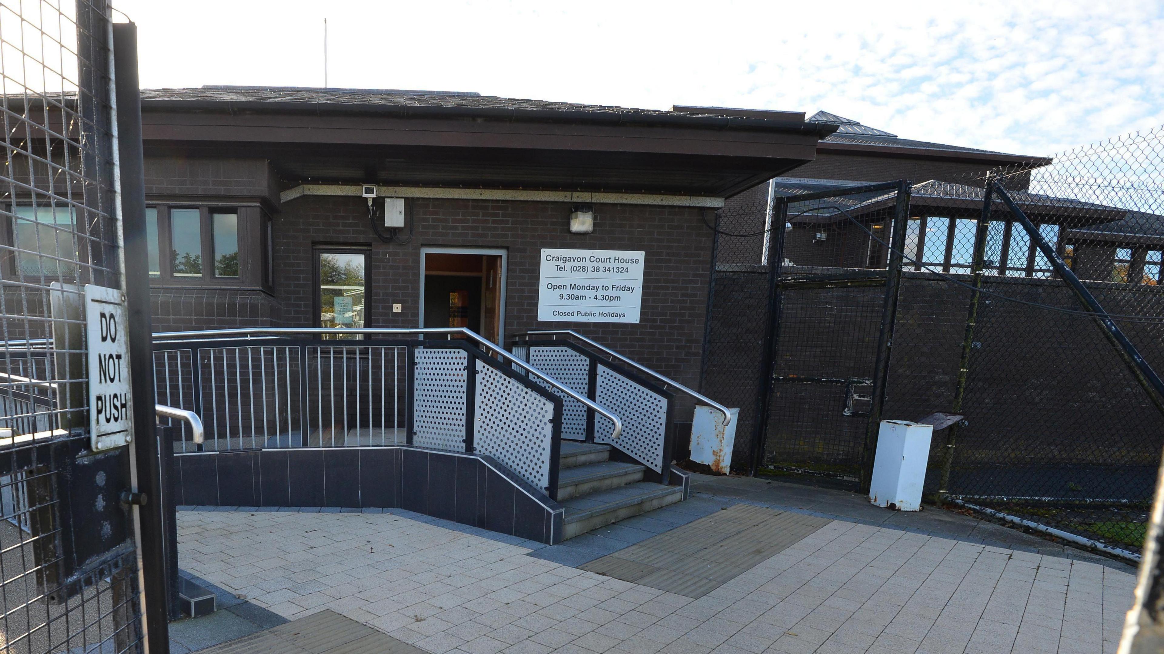 The outside of a brown brick building with high fences around it.  A sign next to the door reads "Craigavon Court House"