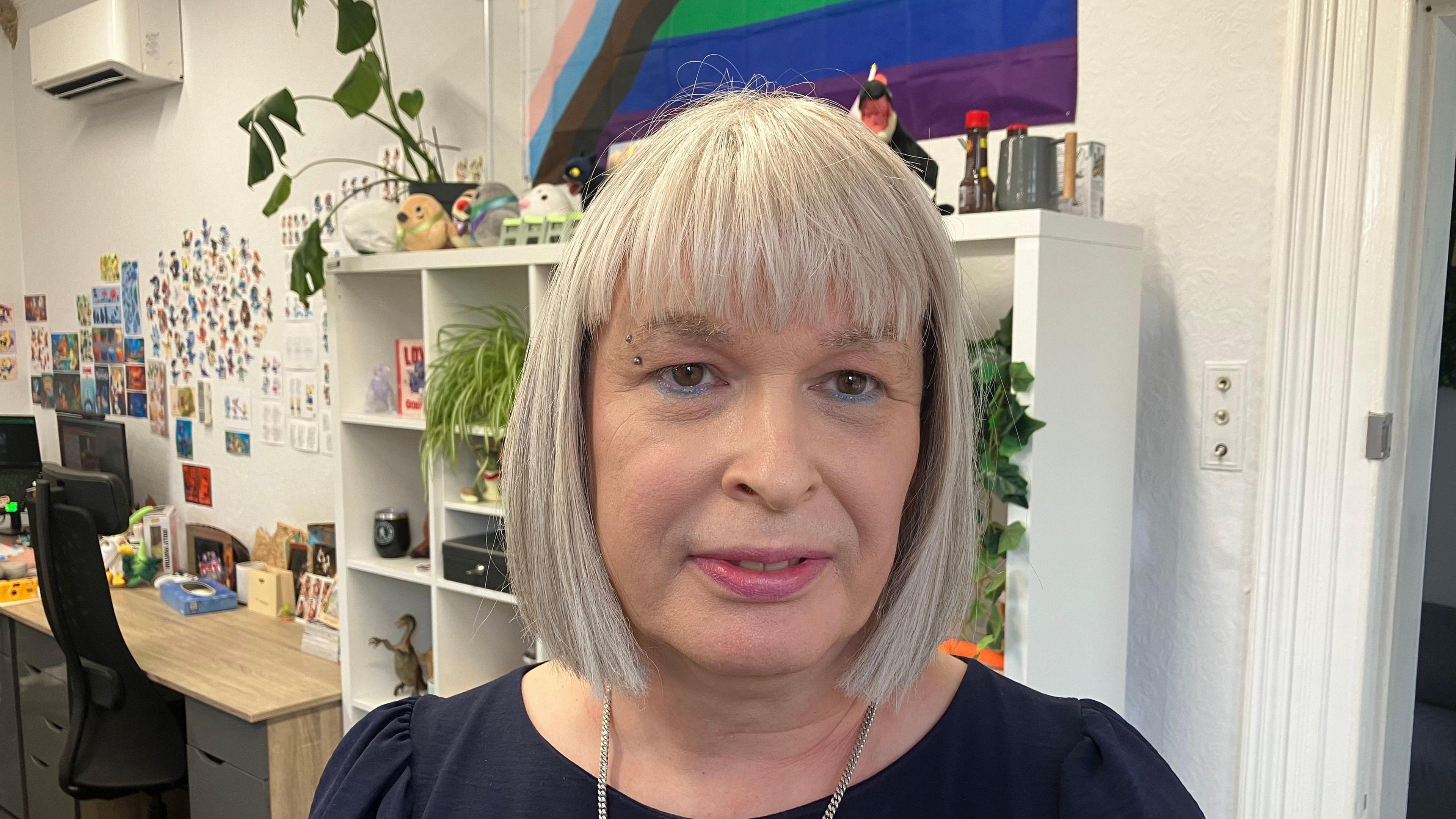 Kerry has a short grey haircut, an eyebrow piercing and blue eyeliner. She is looking directly at the camera and is stood in front of a cabinet which has plants and toys on it. 