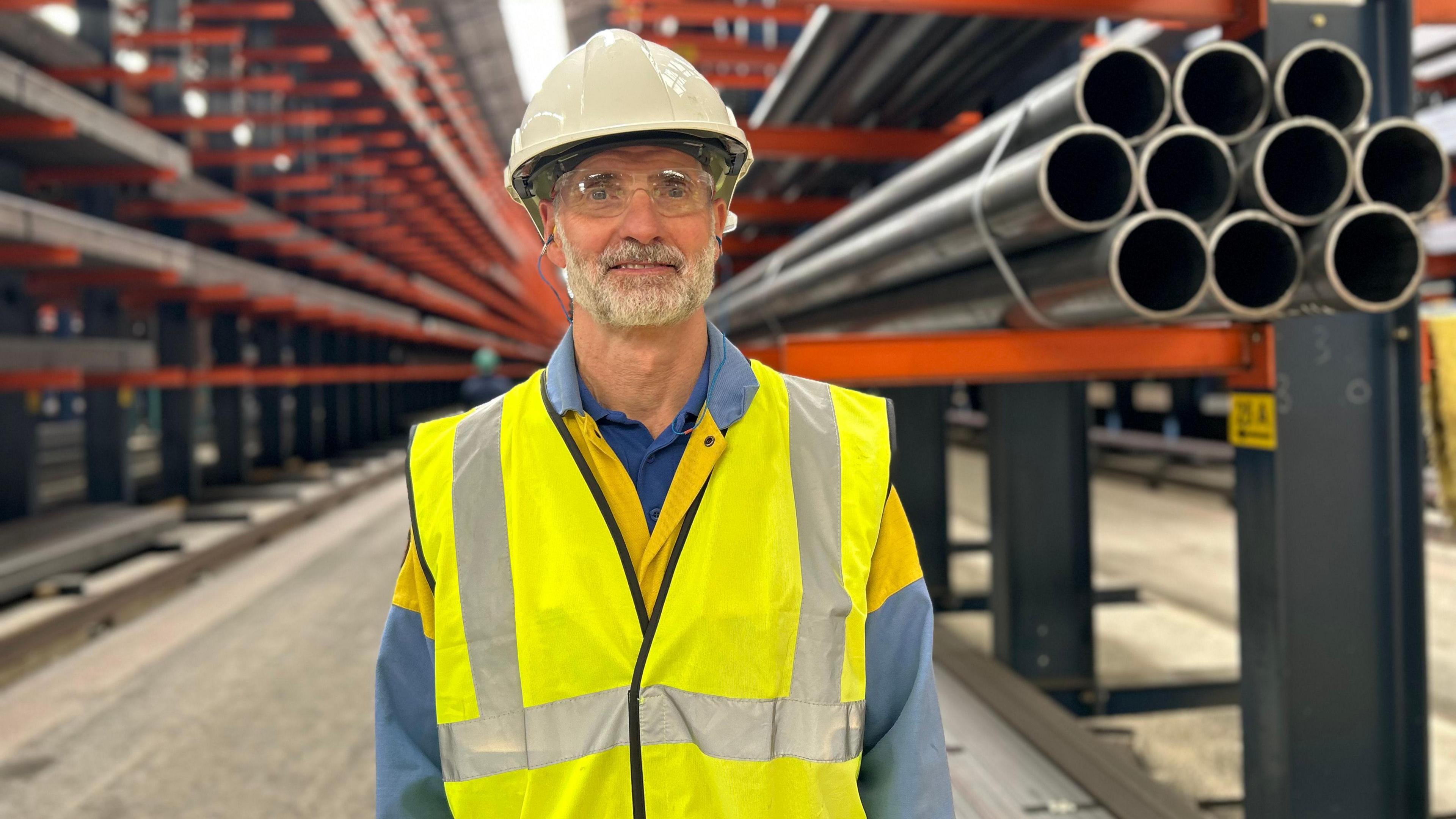 Pual Ilko, who has a white beard and is wearing safety glasses, a high-vis jacket and a white hard hat, stands next to rows of steel tubes 