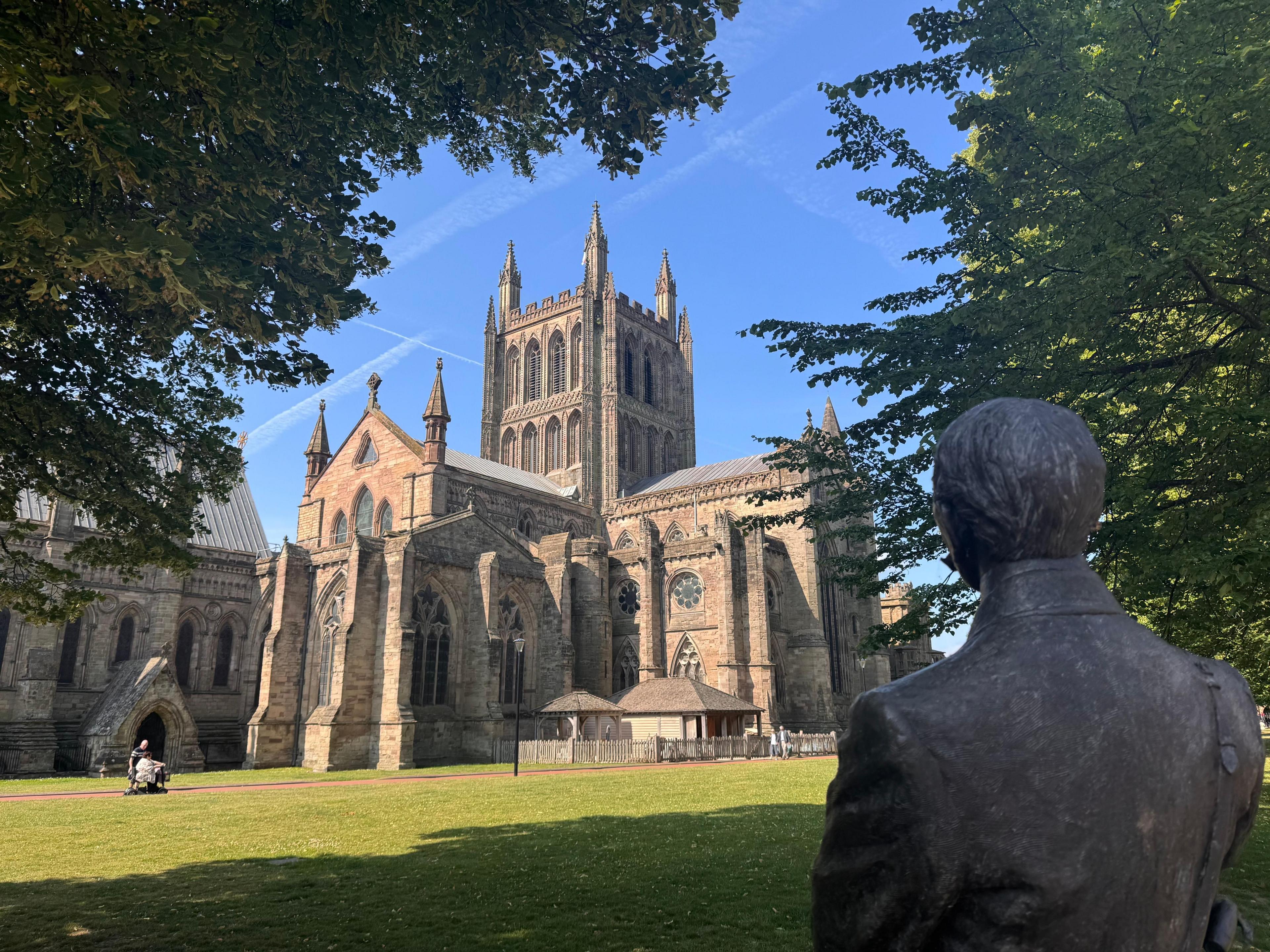 In the foreground, the head and shoulders of a bronze sculpture are shaded, looking away from the camera, through trees, towards a cathedral building. The tower, walls and spires are all in a light brown stone with arched and round windows.  