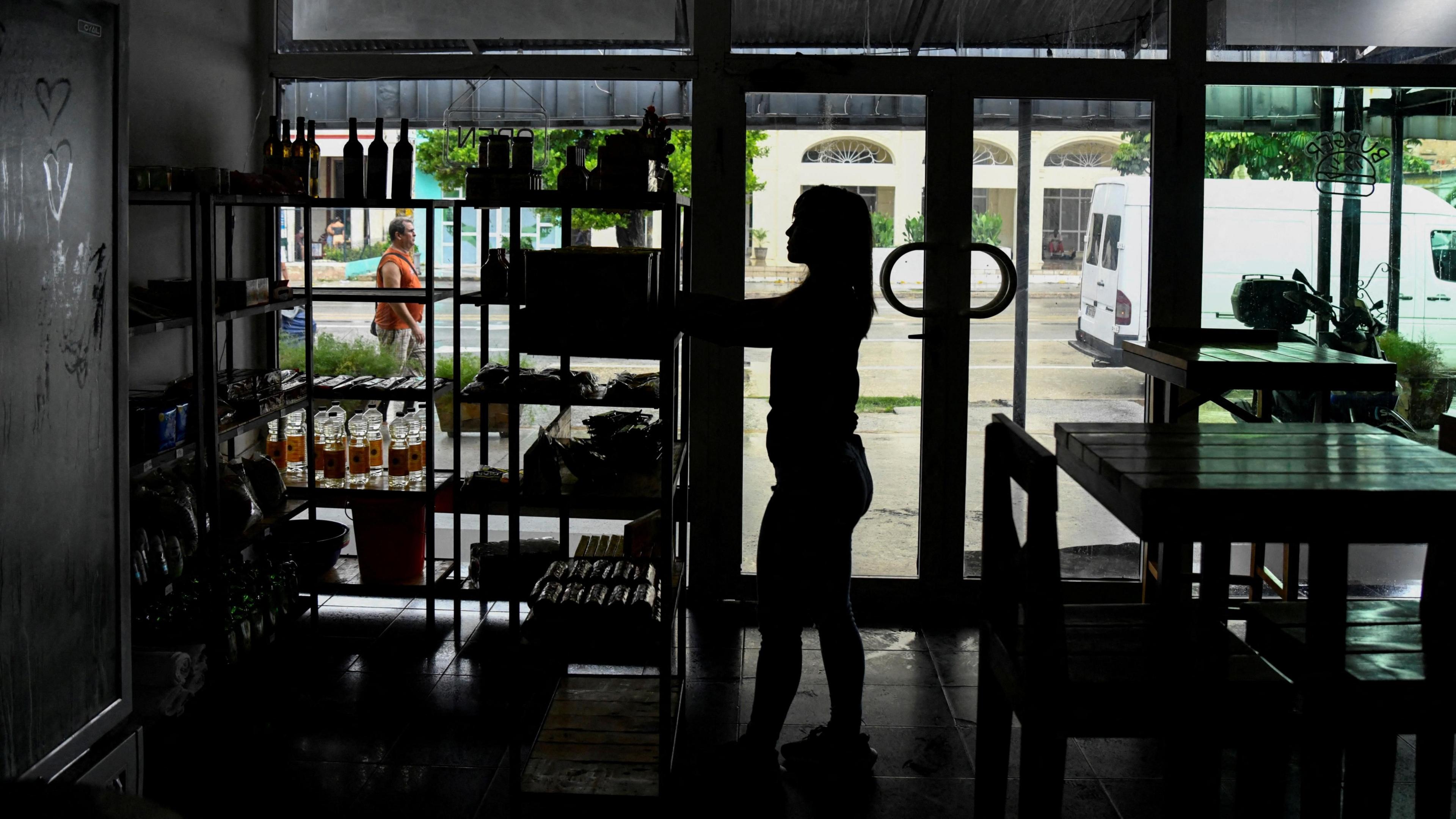 A woman works in a restaurant during a blackout in Havana, Cuba. She's in shadow and is looking at something on a rack of shelves.