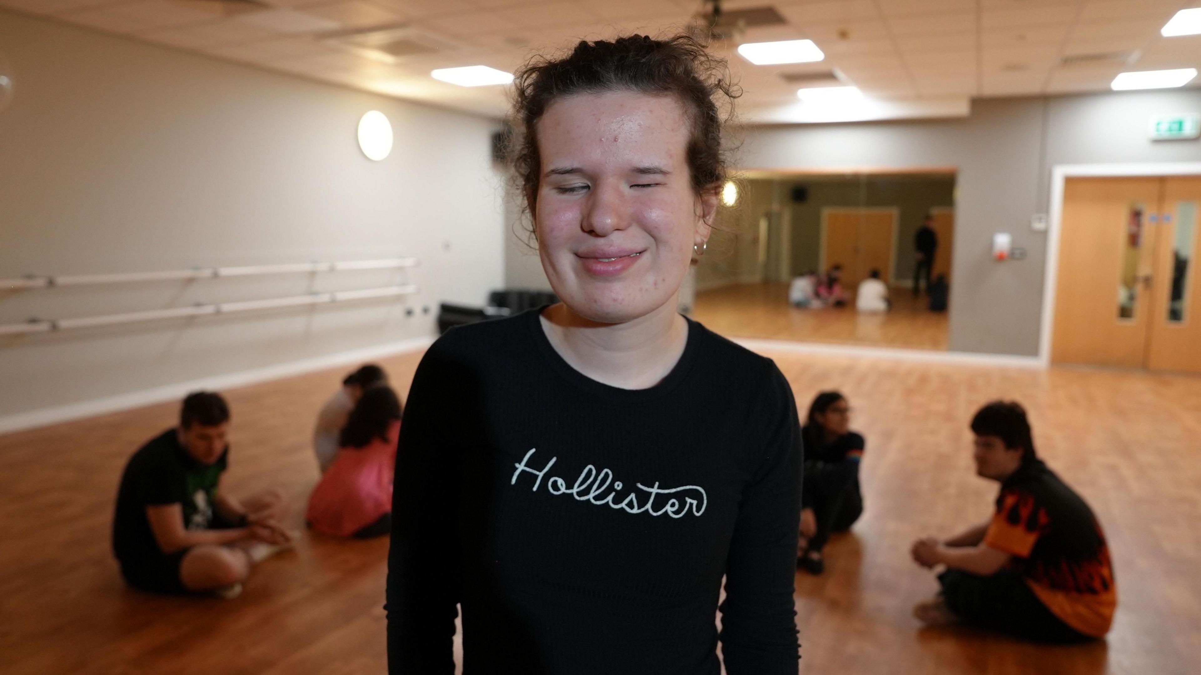 A girl with curly hair and a black Hollister top smiles at the camera, while students sit behind her on the light wood floor of a dance studio. There are bars on the wall to the left and a large mirror at the far end of the room. 