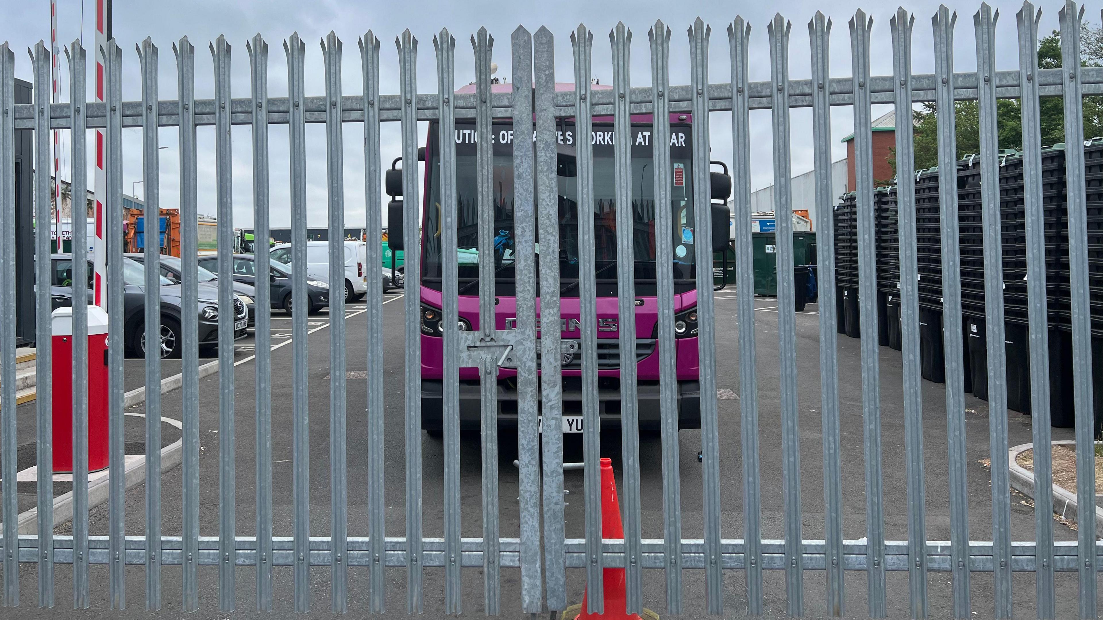 A large purple waste lorry stationary behind a big iron gate that is shut with an orange cone in front of it