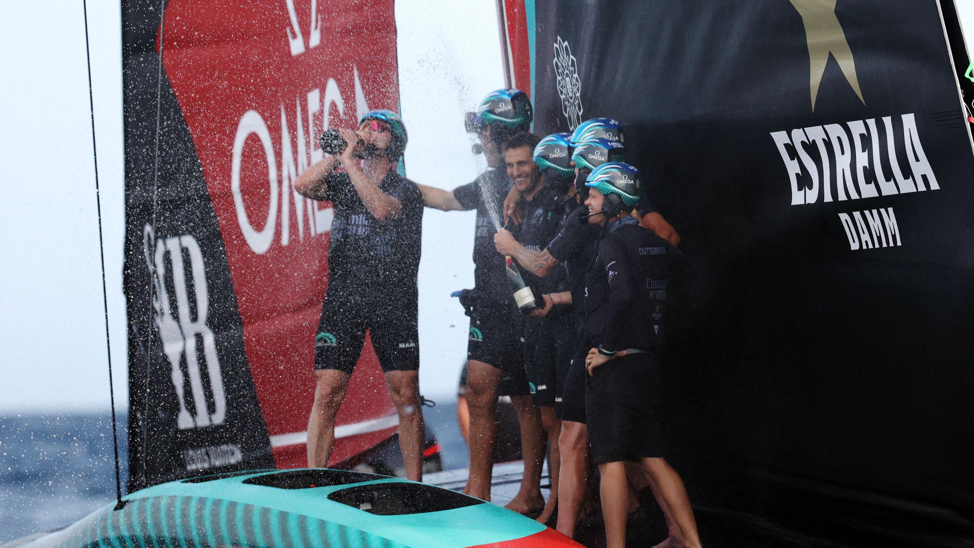 Six men in black wet suits standing on deck of a boat drinking champagne in celebration