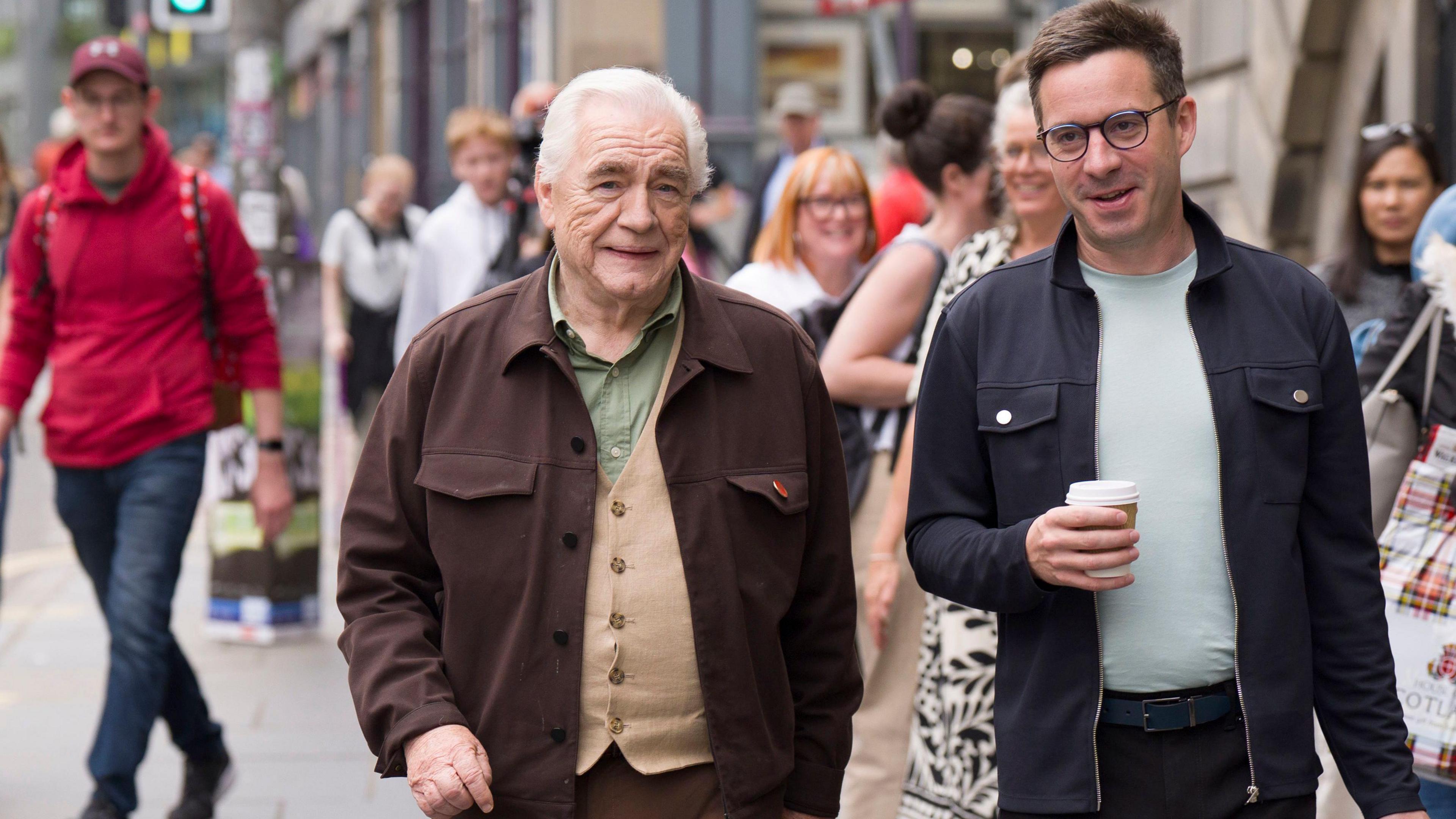 Actor Brian Cox  in green shirt and brown jacket walks along the Royal Mile in Edinburgh with James Graham, a bespectacled man in a blue jacket and green T shirt. He is carrying a disposable drinking cup.