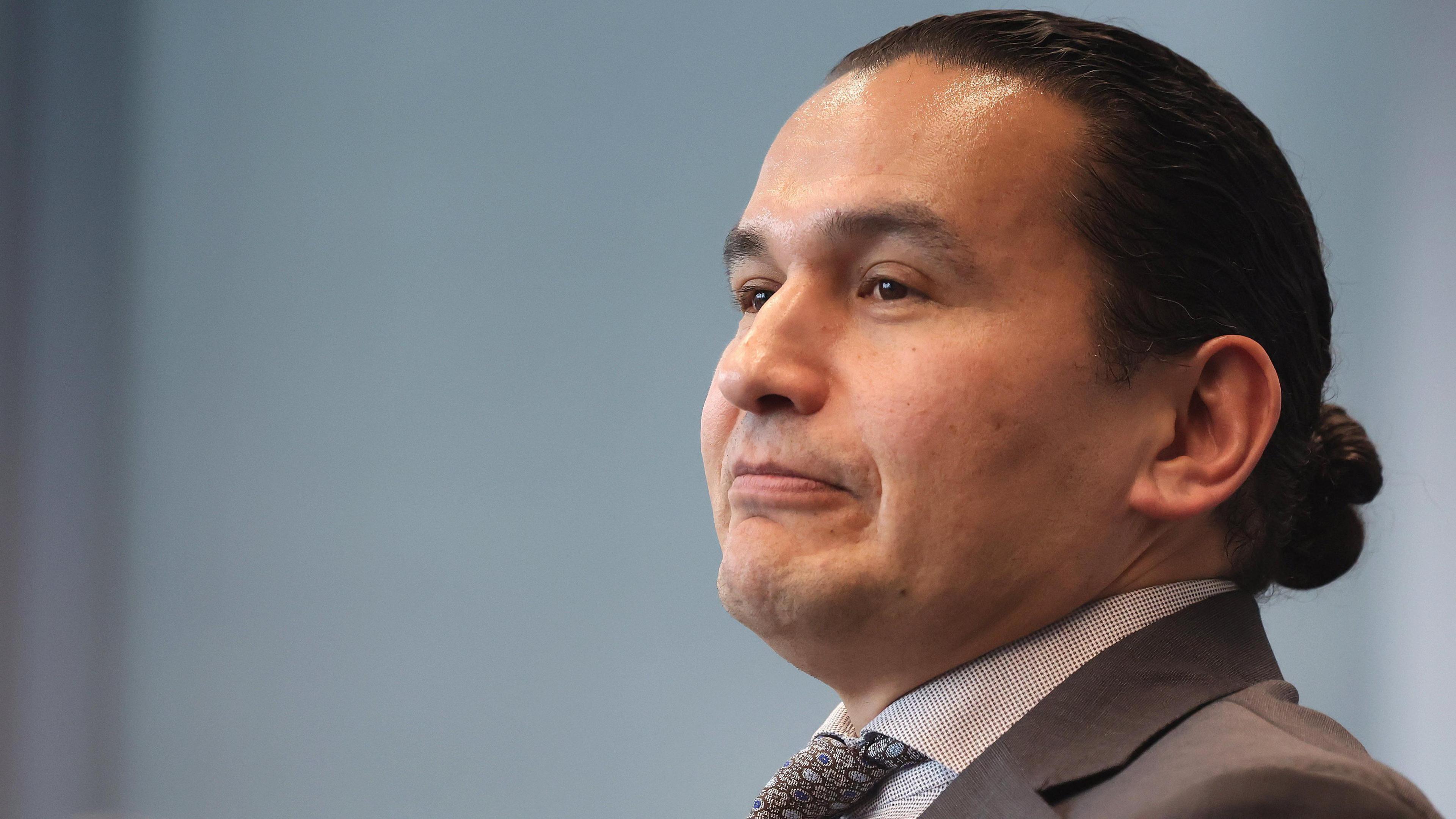 Manitoba Premier Wab Kinew is seen in three-quarter profile with a neutral expression on his face. There is a pale blue non-descript background and he is wearing a grey suit and tie. 
