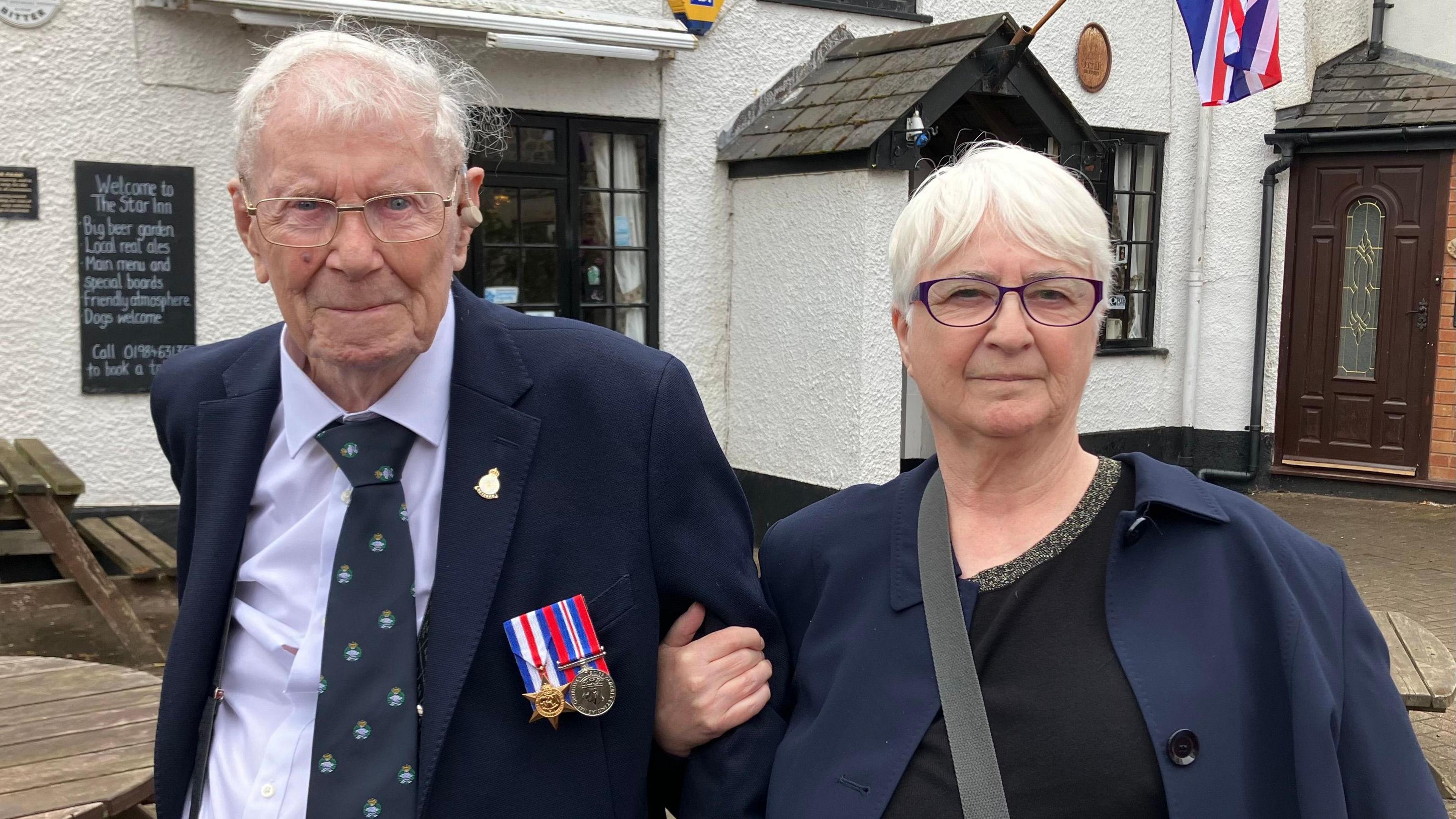 George Lewis and Liz Lewis standing outside a pub. They both have white hair and wear glasses, and Liz is holding her father's arm. George wears a dark suit and tie with his two medals clearly on display, and Liz wears a black top and navy jacket. 