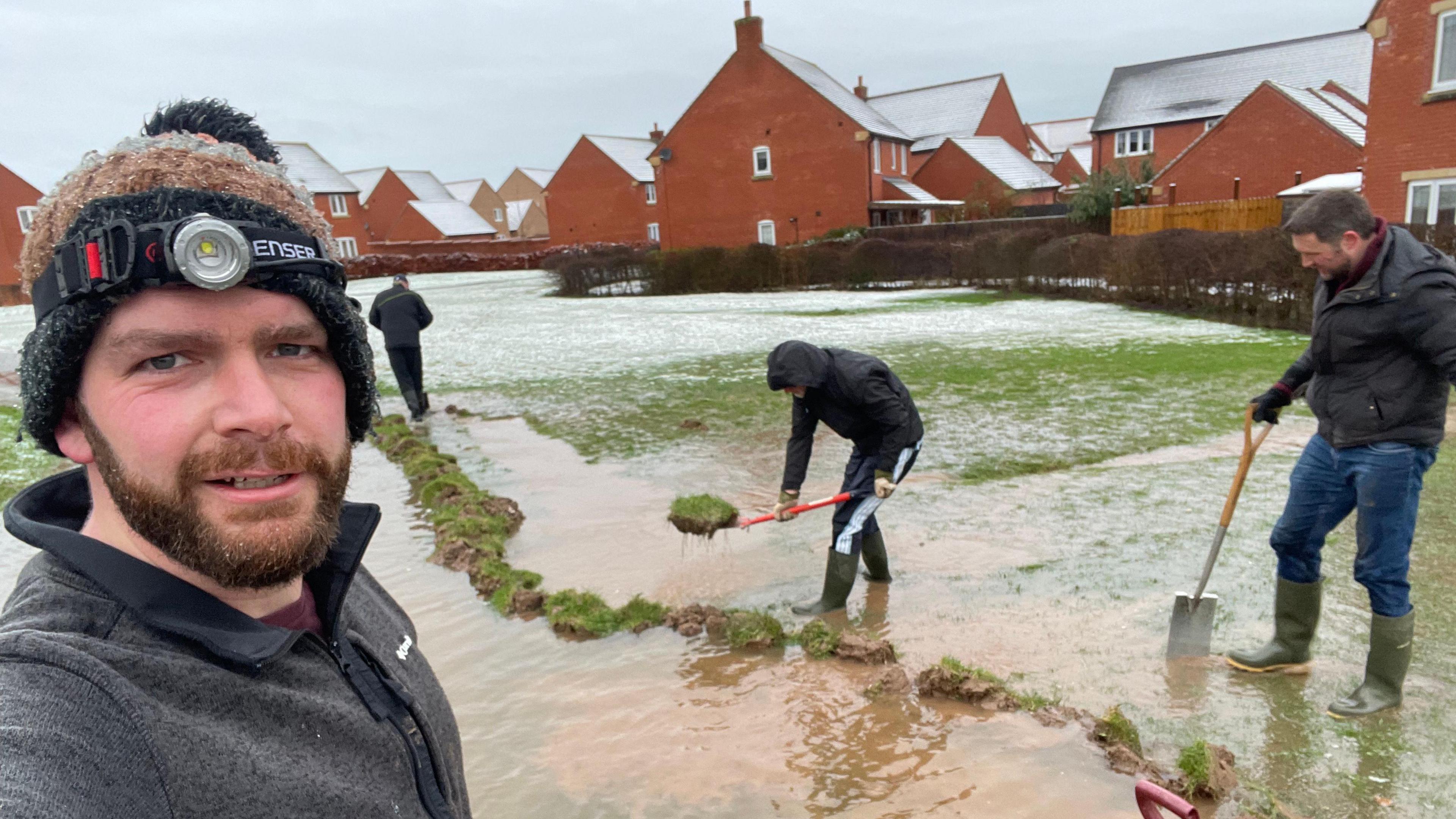 Resident Ben Stockley is pictured with residents armed with garden tools as they dig trenches in a field alongside a stream of floodwater. There is a housing estate behind them. 