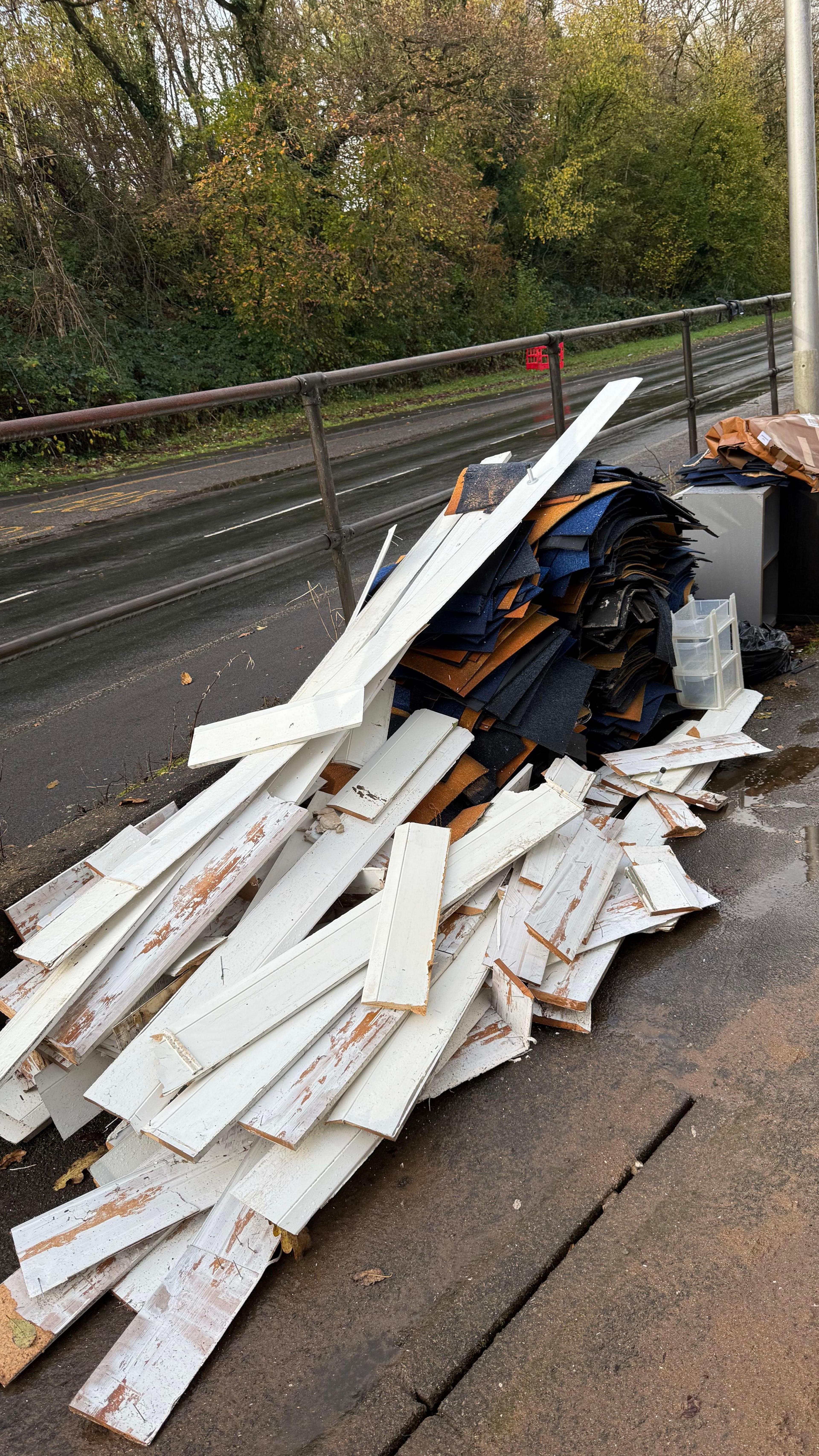 Piles of white skirting boards and carpet piled up on the pavement next to a road. Dirt, debris and some water on the ground and in the background there are some trees with green and yellow leaves. 