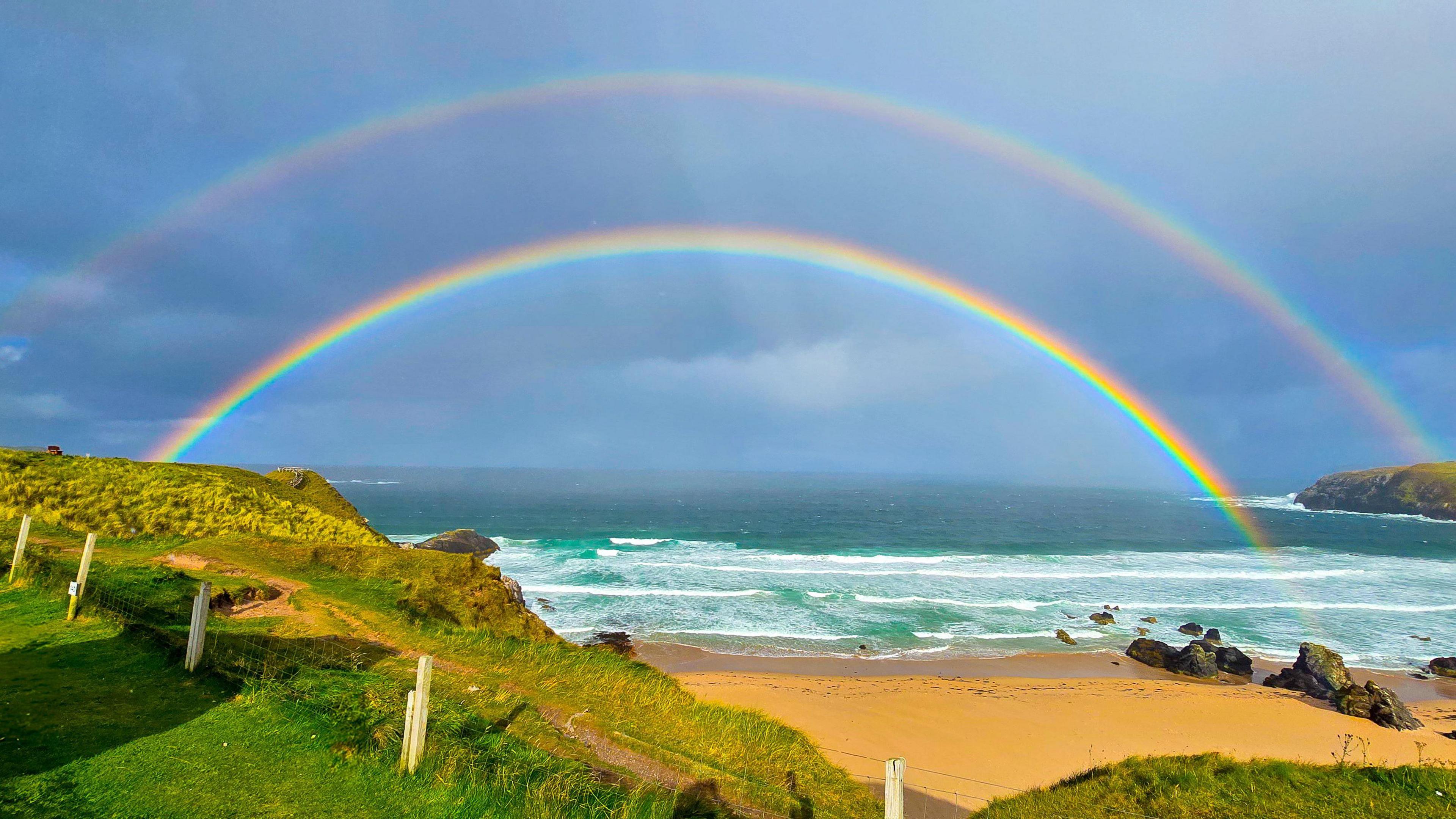 A double rainbow in the sky over a cloudy Durness Beach. The larger rainbow is slightly more faded. The smaller one is much brighter. Both are over water which has a slight green tint, with white waves rolling on to a dark golden beach, with a few rocks on the sand. In the foreground, there is green grass and small cliffs covered with green plants and white fence posts.