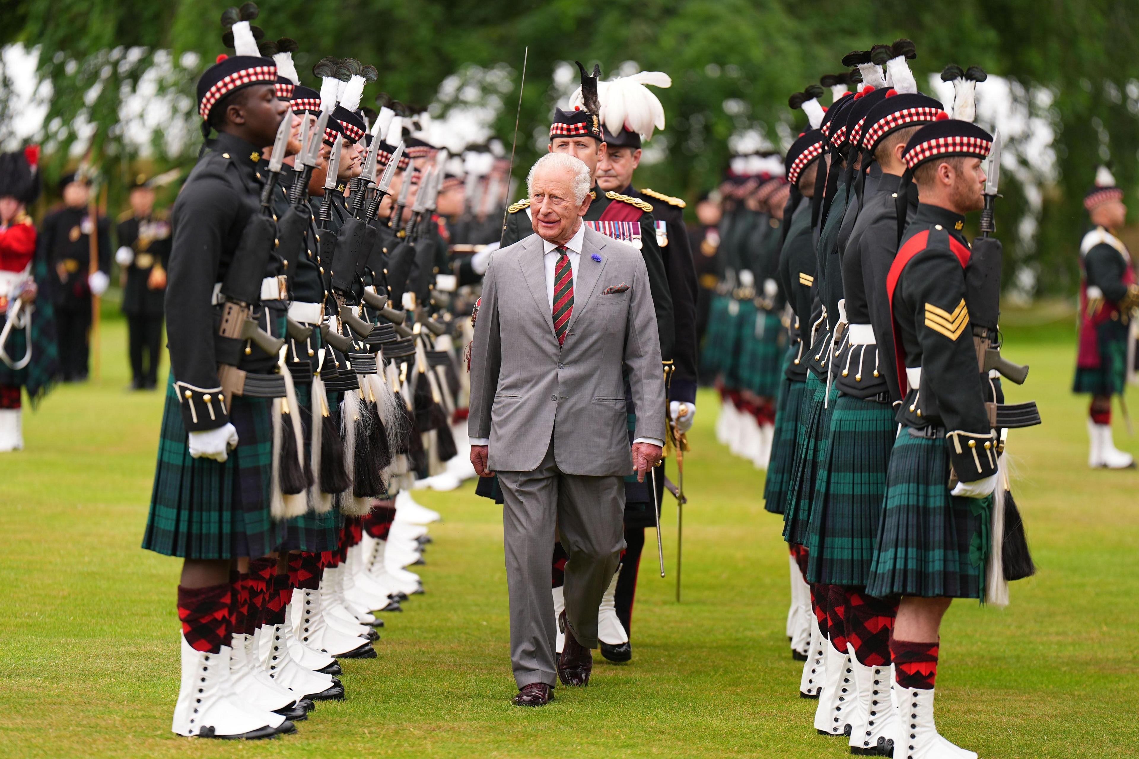 King Charles III wearing a grey suit with a white shirt and red and green striped tie. He is walking through the Palace Guard - a line of soldiers, wearing black uniforms and green tartan kilts.
