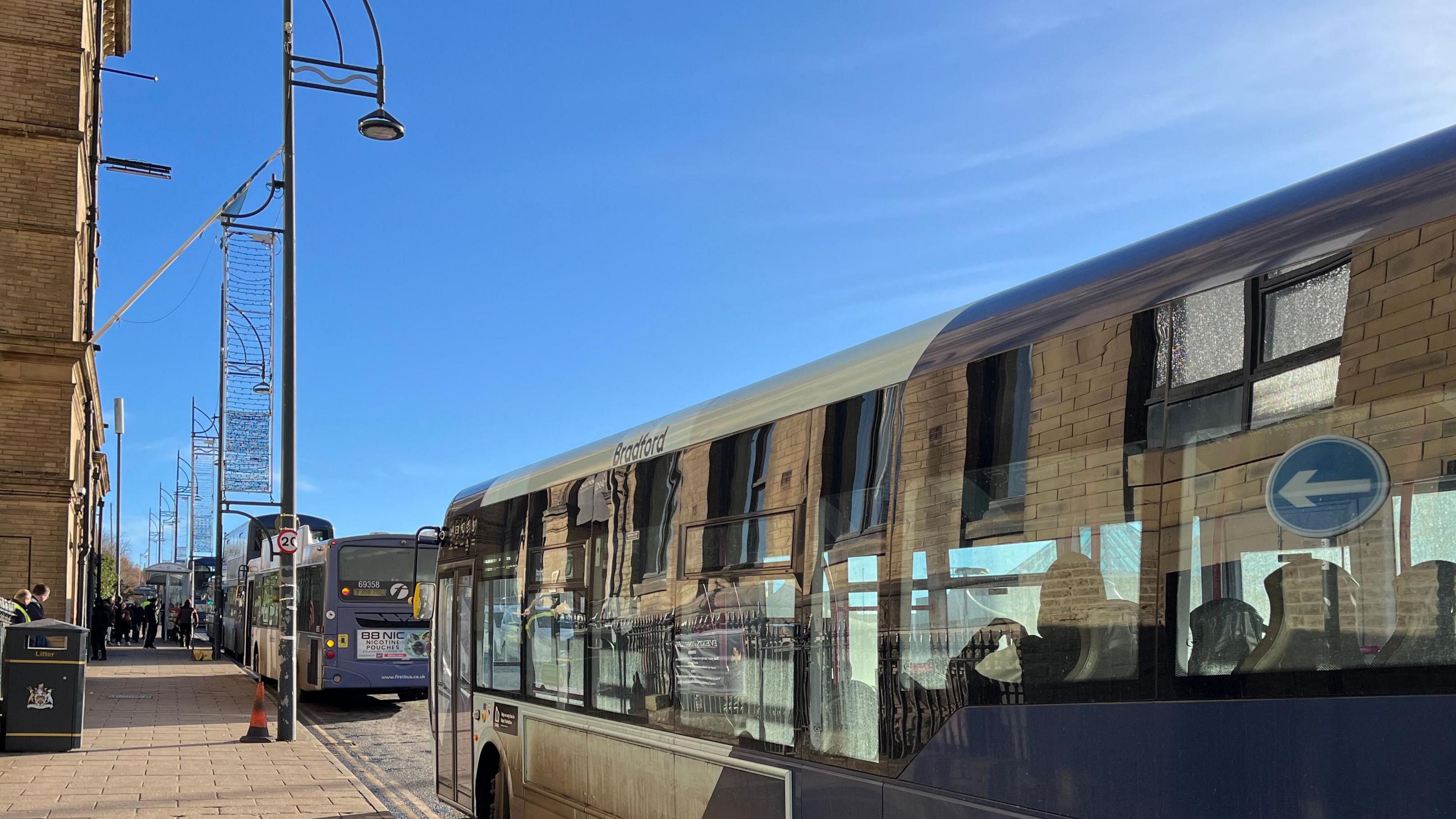 A number of buses along a road with a pavement to the left of the image