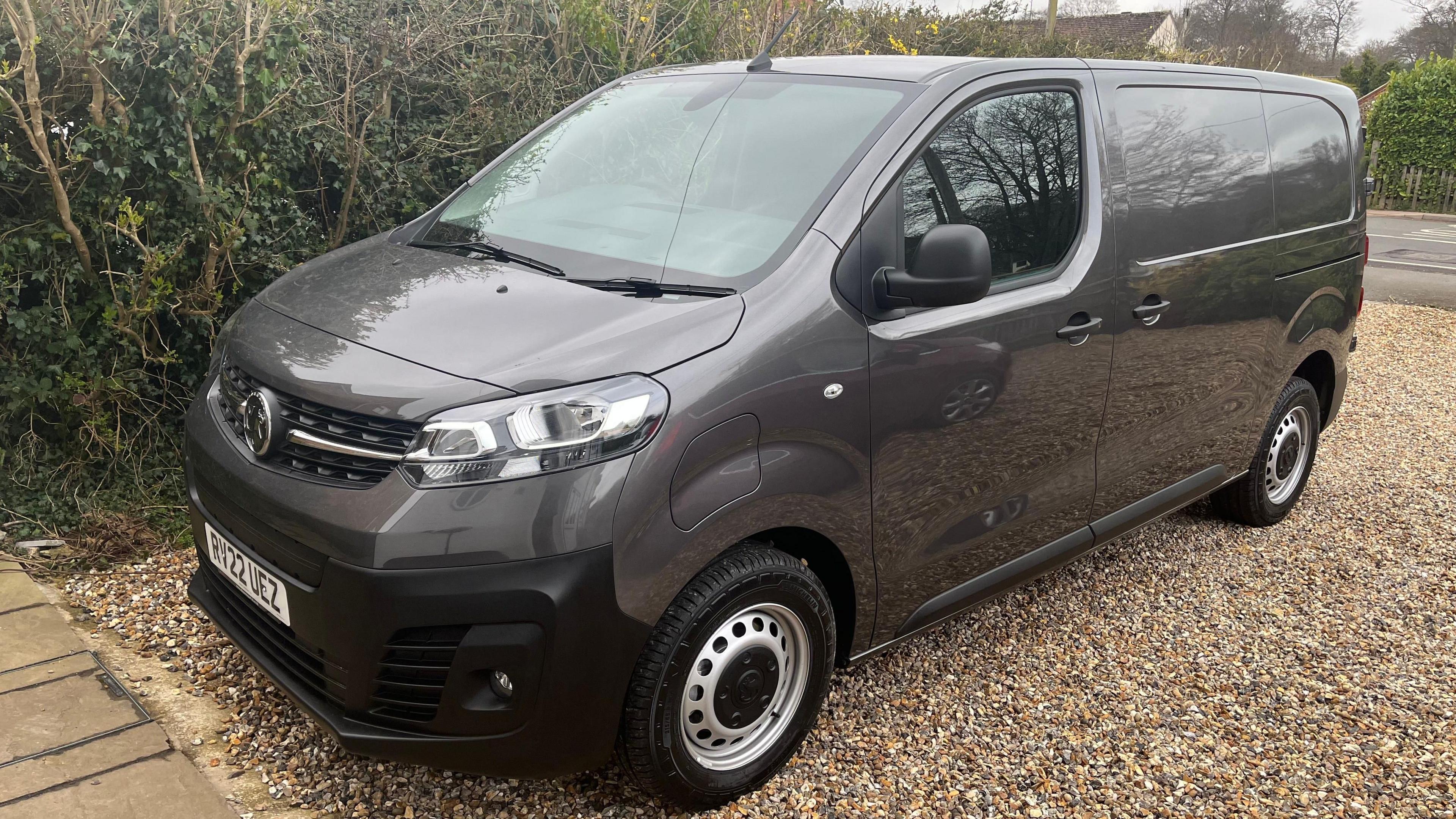 A grey Vauxhall commercial van is parked on a gravel driveway near hedges, with a UK number plate visible.