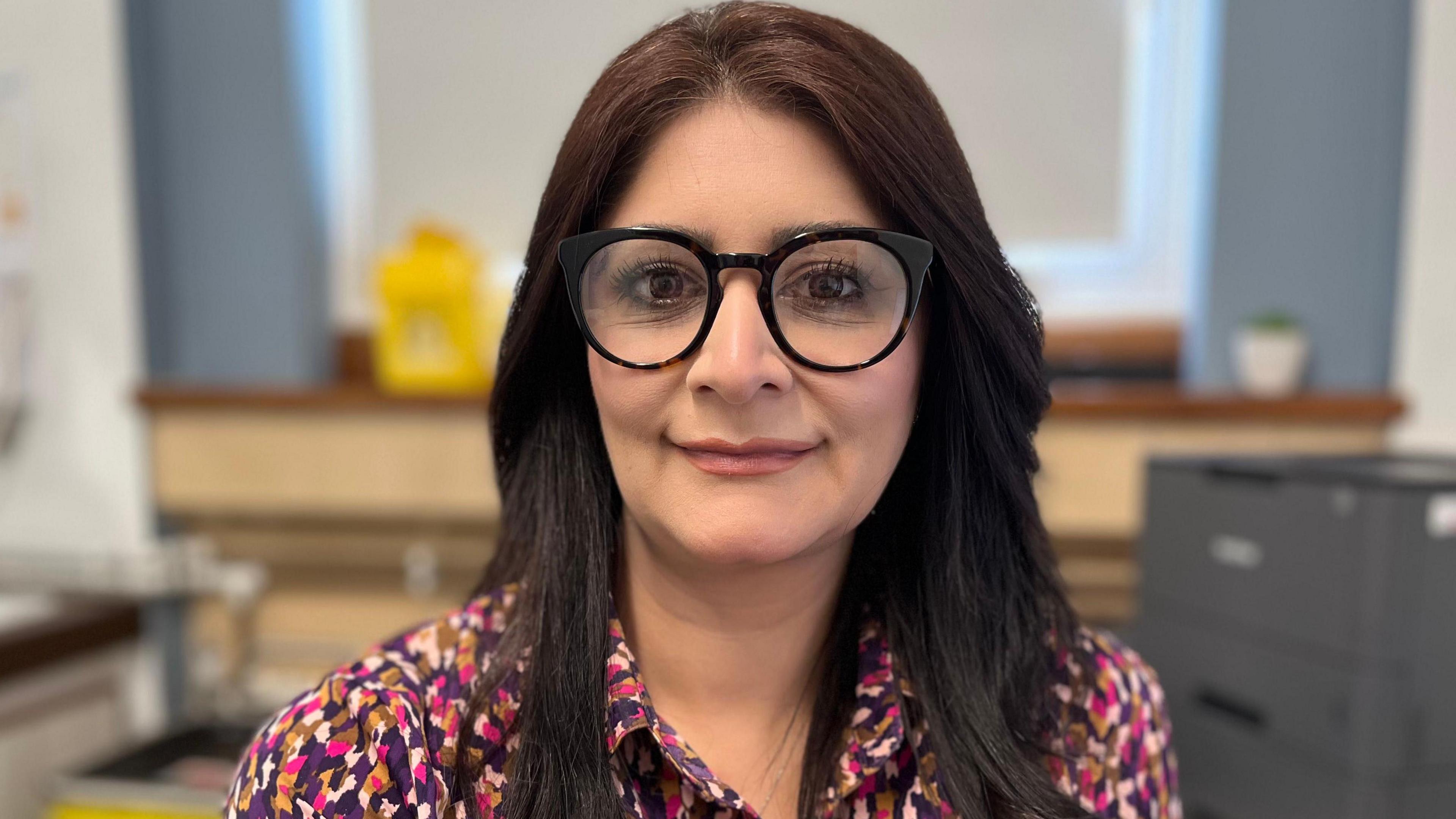 Dr Qayyum has long dark hair and is smiling at the camera. She is wearing black round framed glasses and a purple, pink and red patterned blouse. 
