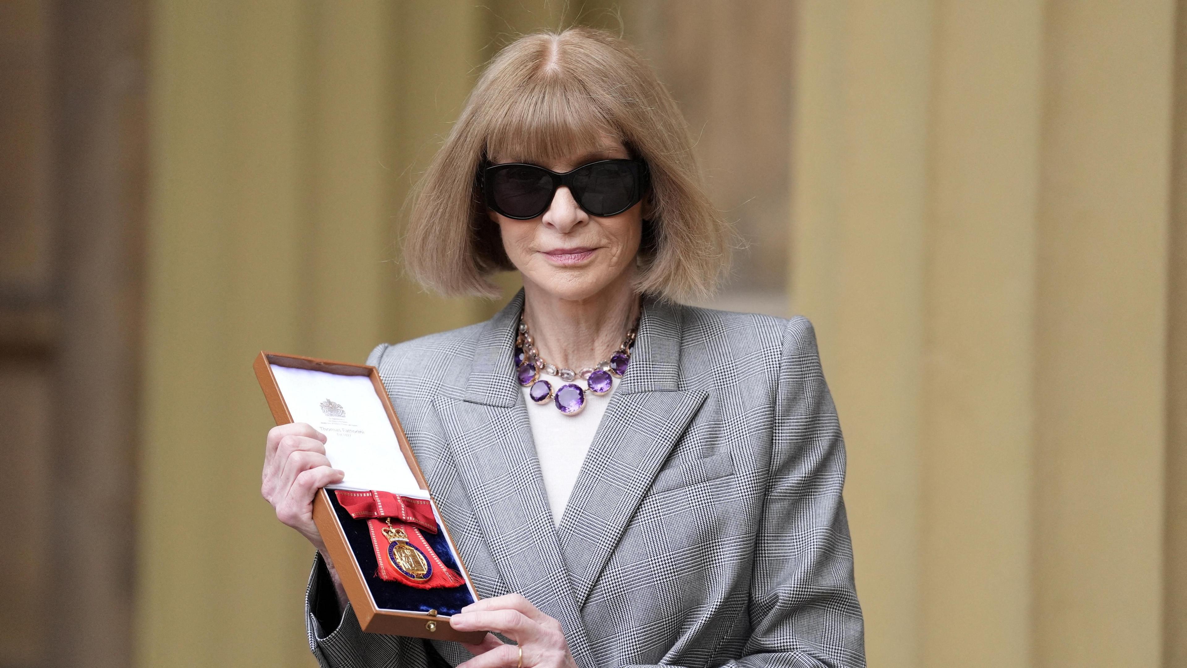 Anna Wintour holds her Companion of Honour medal outside Buckingham Palace, wearing a grey blazer and amethyst necklace. Her sunglasses are firmly on. 