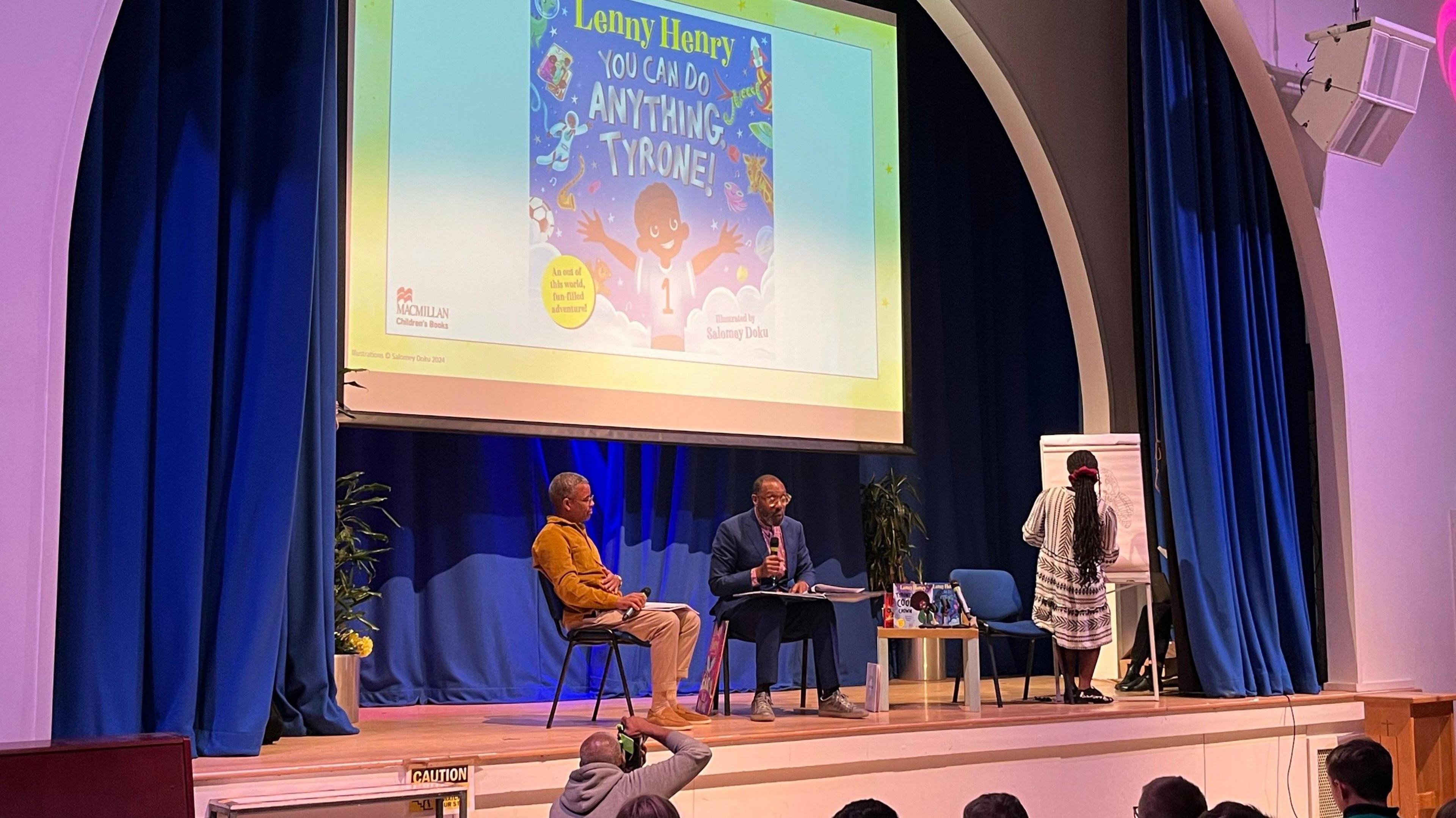 Sir Lenny Henry on stage reading his book to school children. The front cover of his book is on a big screen behind him