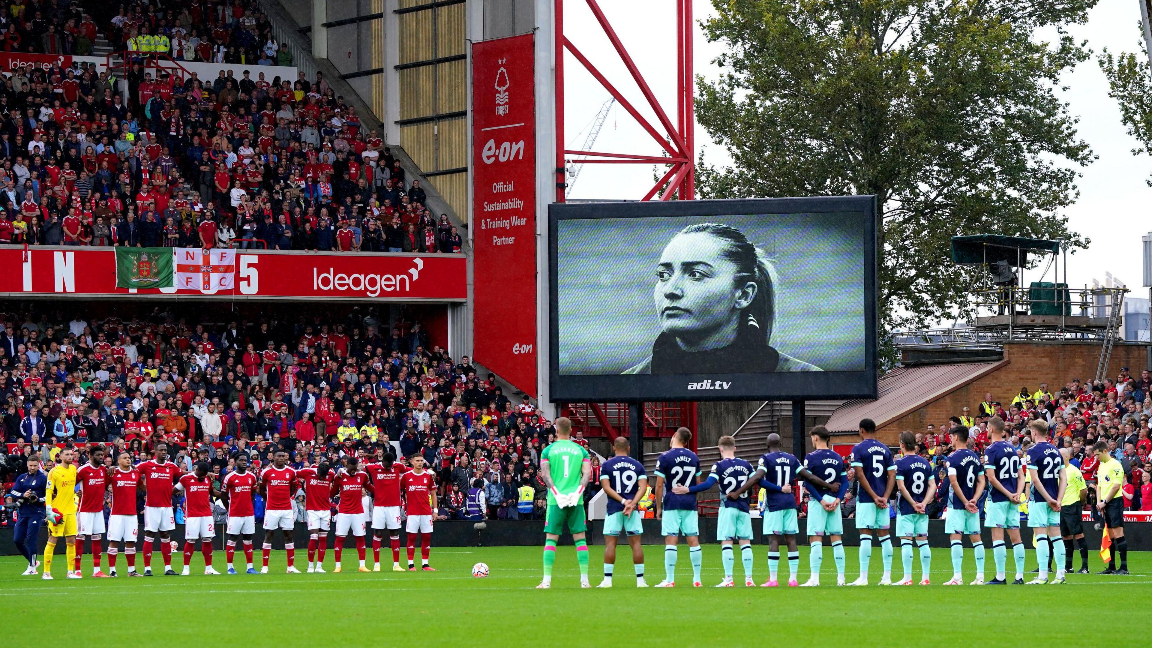 Two rows of football players with their arms around each other with a black and white picture of Maddy Cusack on a large screen at a full stadium