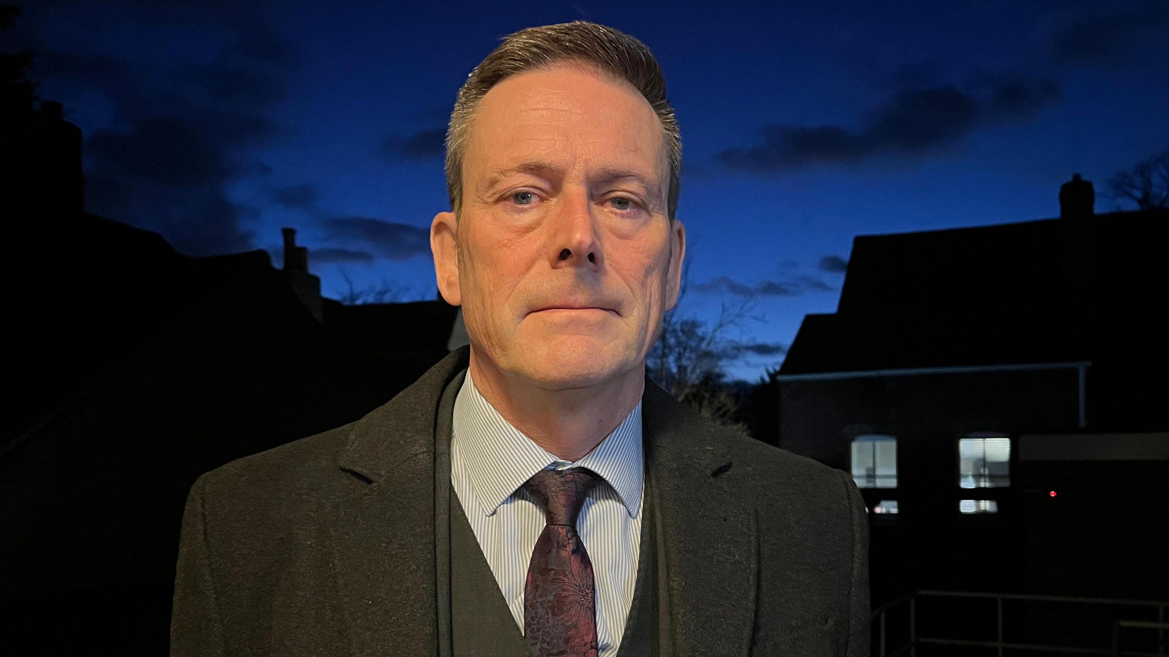 Nick Bell, standing outside in the dark, with houses behind him, and a dark sky. He is looking straight at the camera, with short hair, wearing a suit, shirt, tie, and coat. He looks serious. 