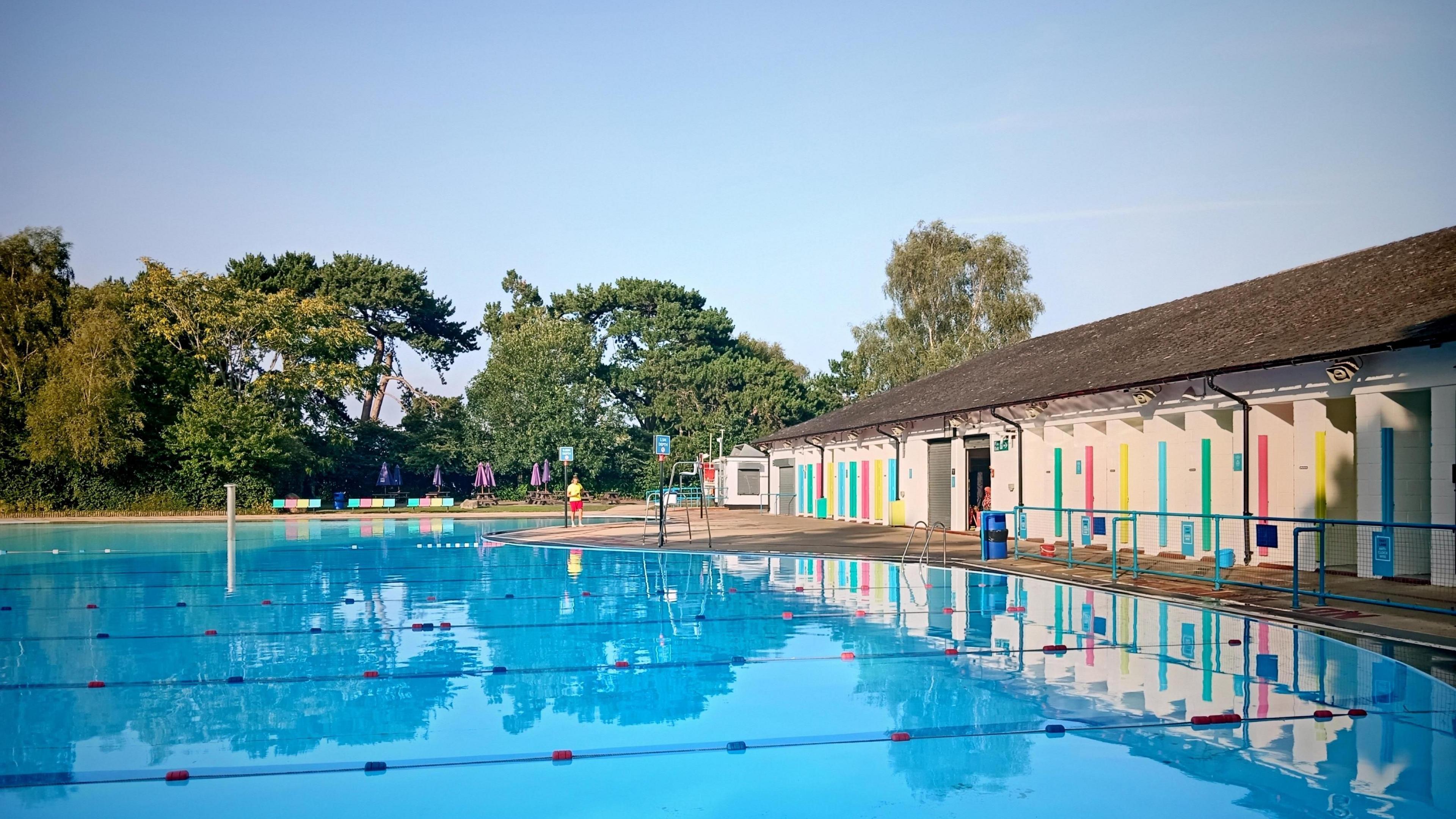 An outdoor swimming pool with lane markers. The blue water is completely still with trees reflected in the water. There is a low white building with colourful doors. On the far side of the pool are trees and a lifeguard wearing a yellow tshirt and red shorts.