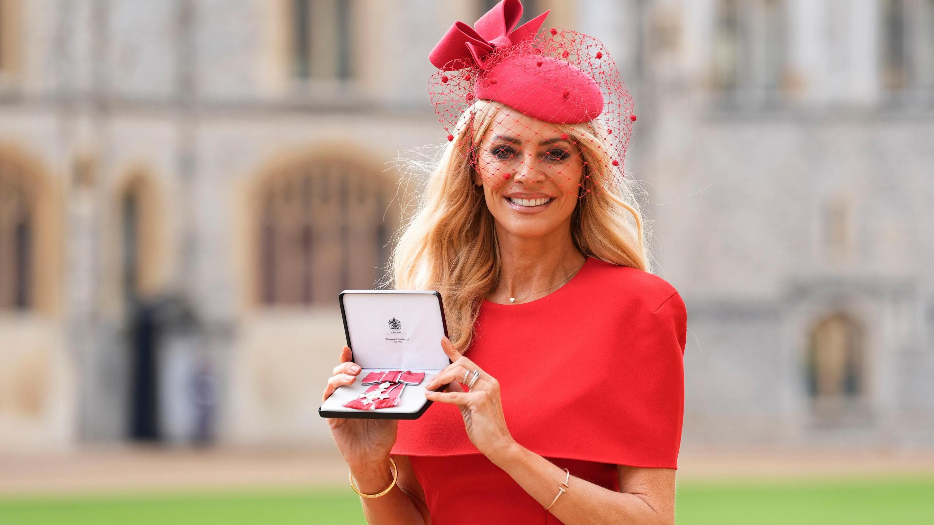 Tess Daly holds her medal after being made a Member of the Order of the British Empire (MBE) at an investiture ceremony at Windsor Castle. She wears a red dress and a fascinator. 