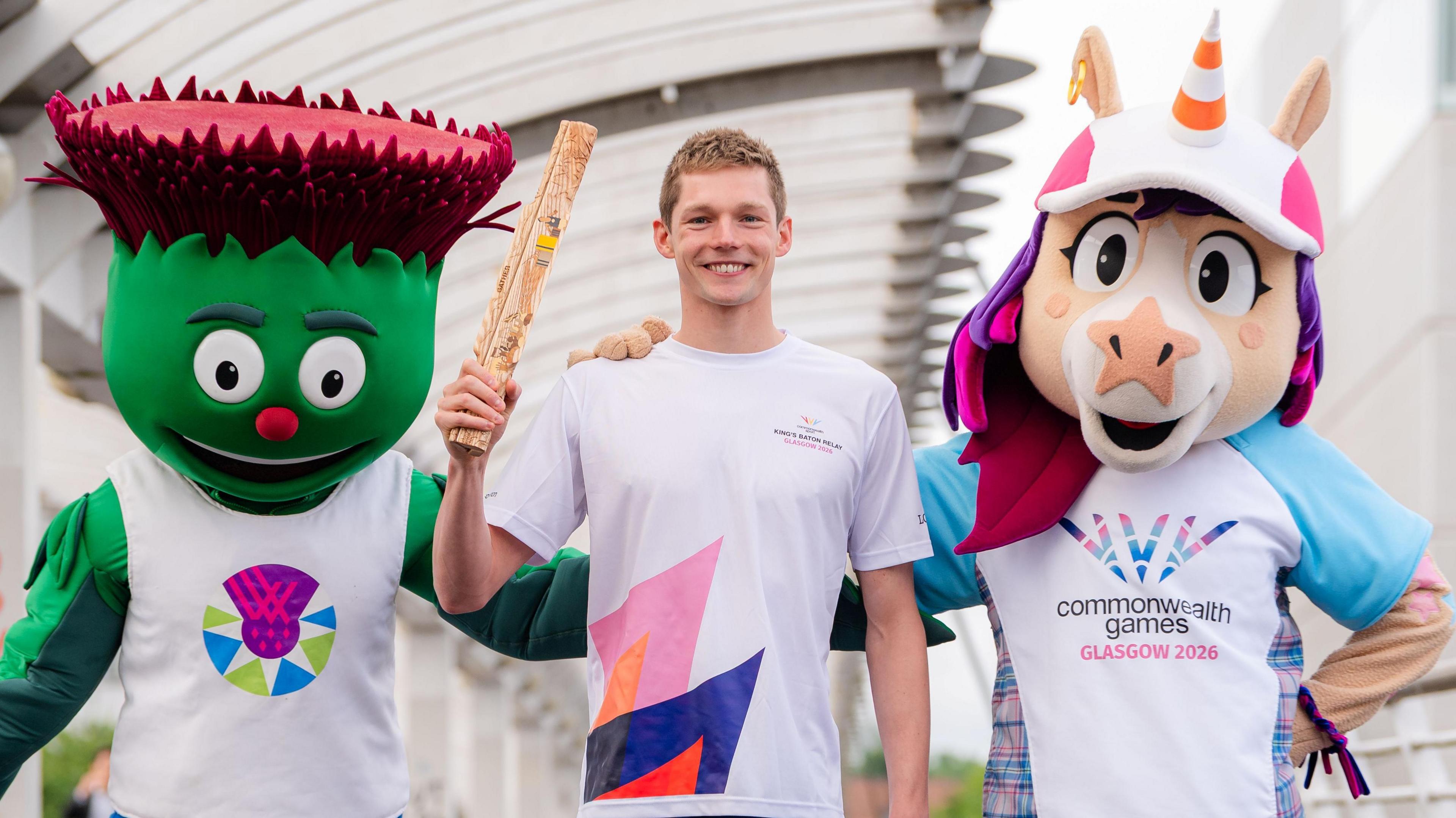 Duncan Scott (centre) holding aloft a wooden Commonwealth Games baton. He is flanked by two mascots - Clyde, a green thistle, and a unicorn called Finnie, who has a traffic cone instead of a conventional horn.  