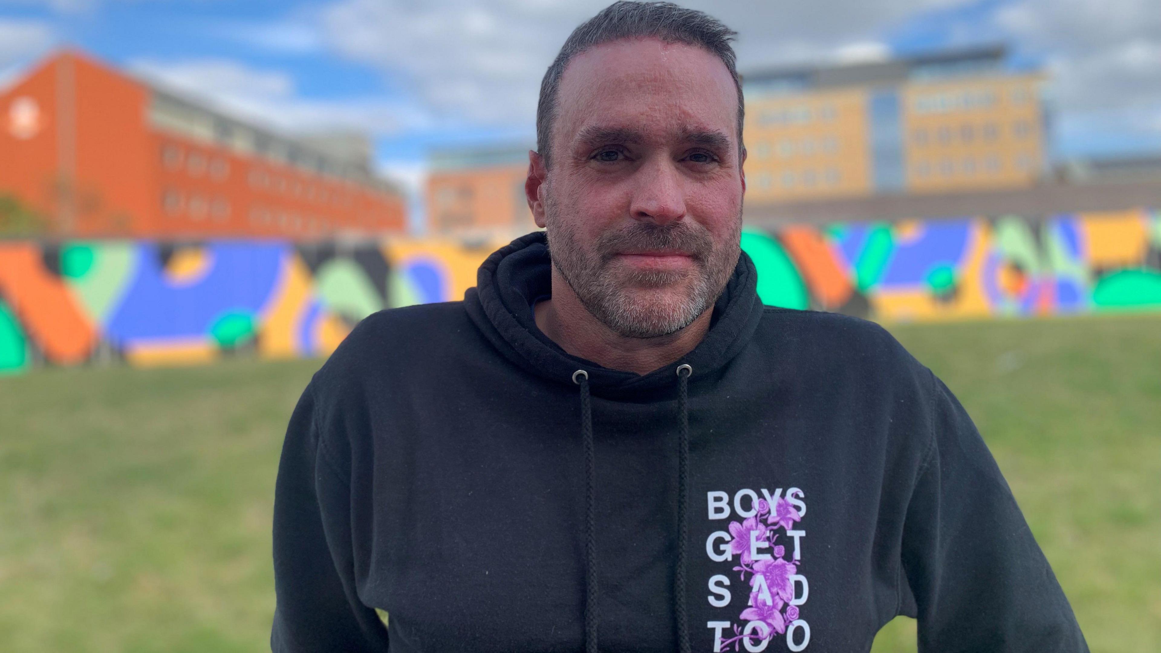 Mr Taylor wearing a black hoodie which has the logo 'Boys get sad too" entwined with purple flowers. He is looking pensive. There is grass in the background and blurry, colourful buildings. 