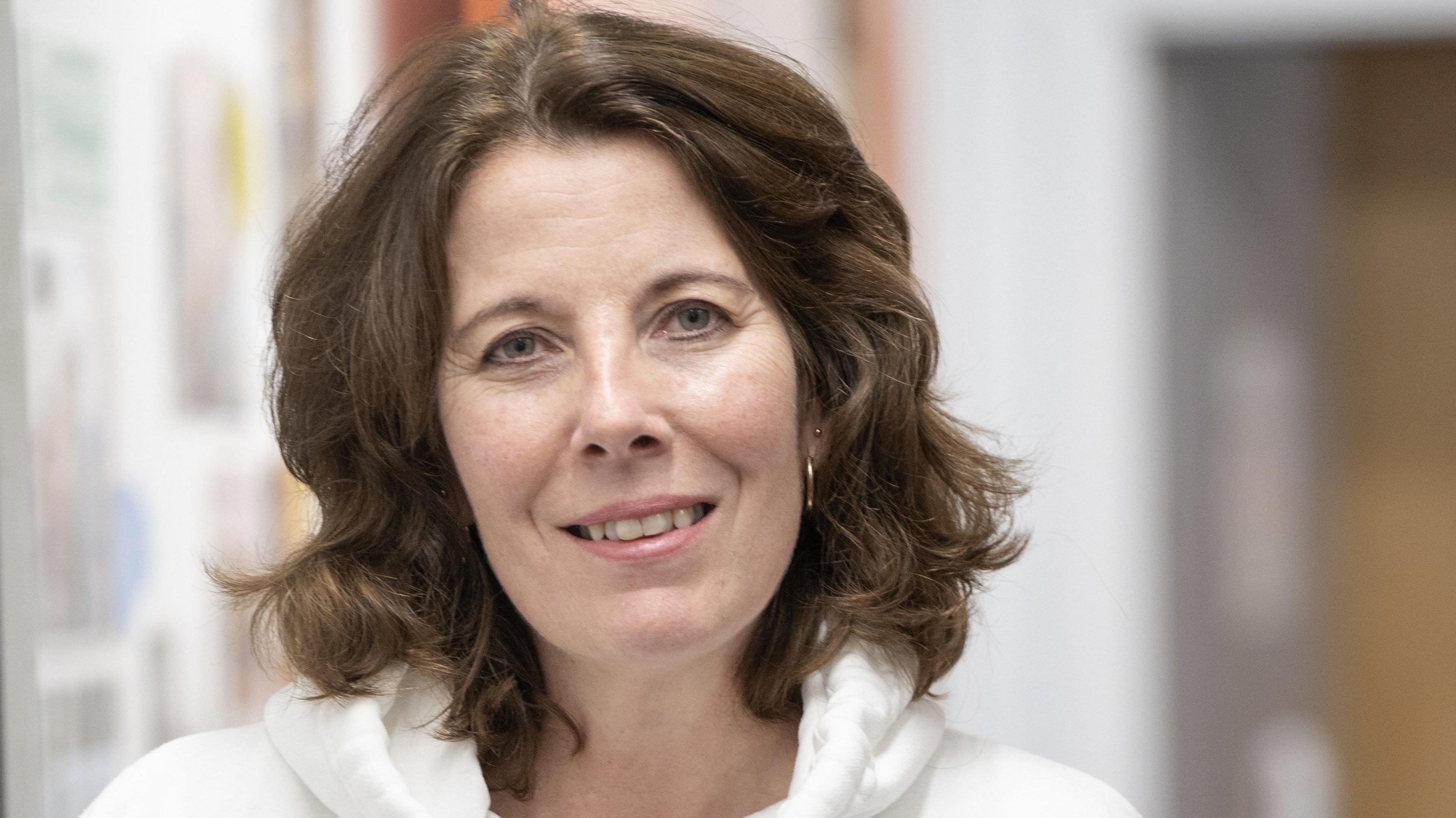 Ms Praill, a woman with dark curly shoulder-length hair and wearing a white hoodie, smiling at the camera.