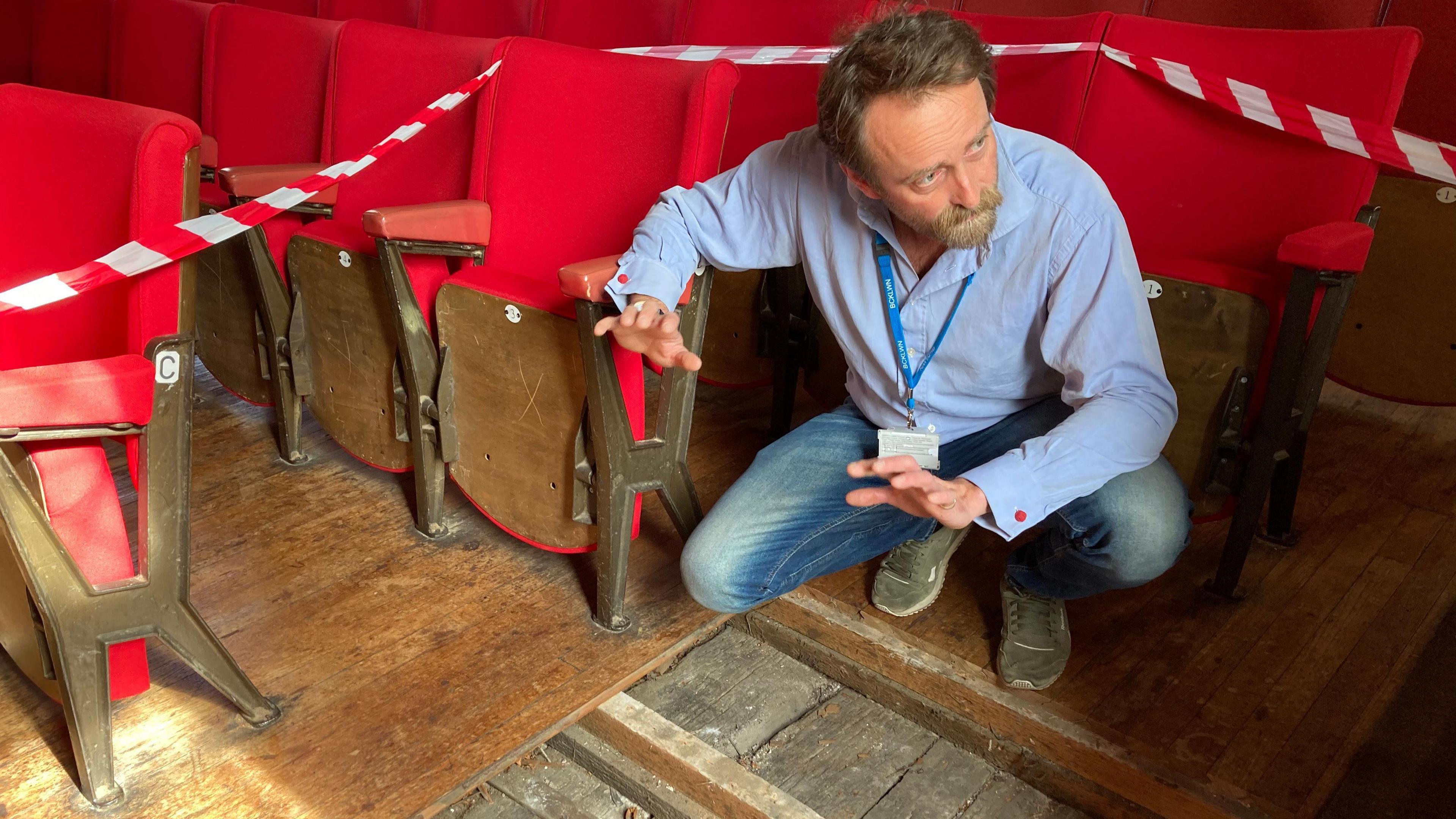 An older picture of Tim FitzHigham before the flooring was fully uncovered. Instead, a small parcel of it has been exposed and Tim is crouching above it. He is surrounded by red seating.